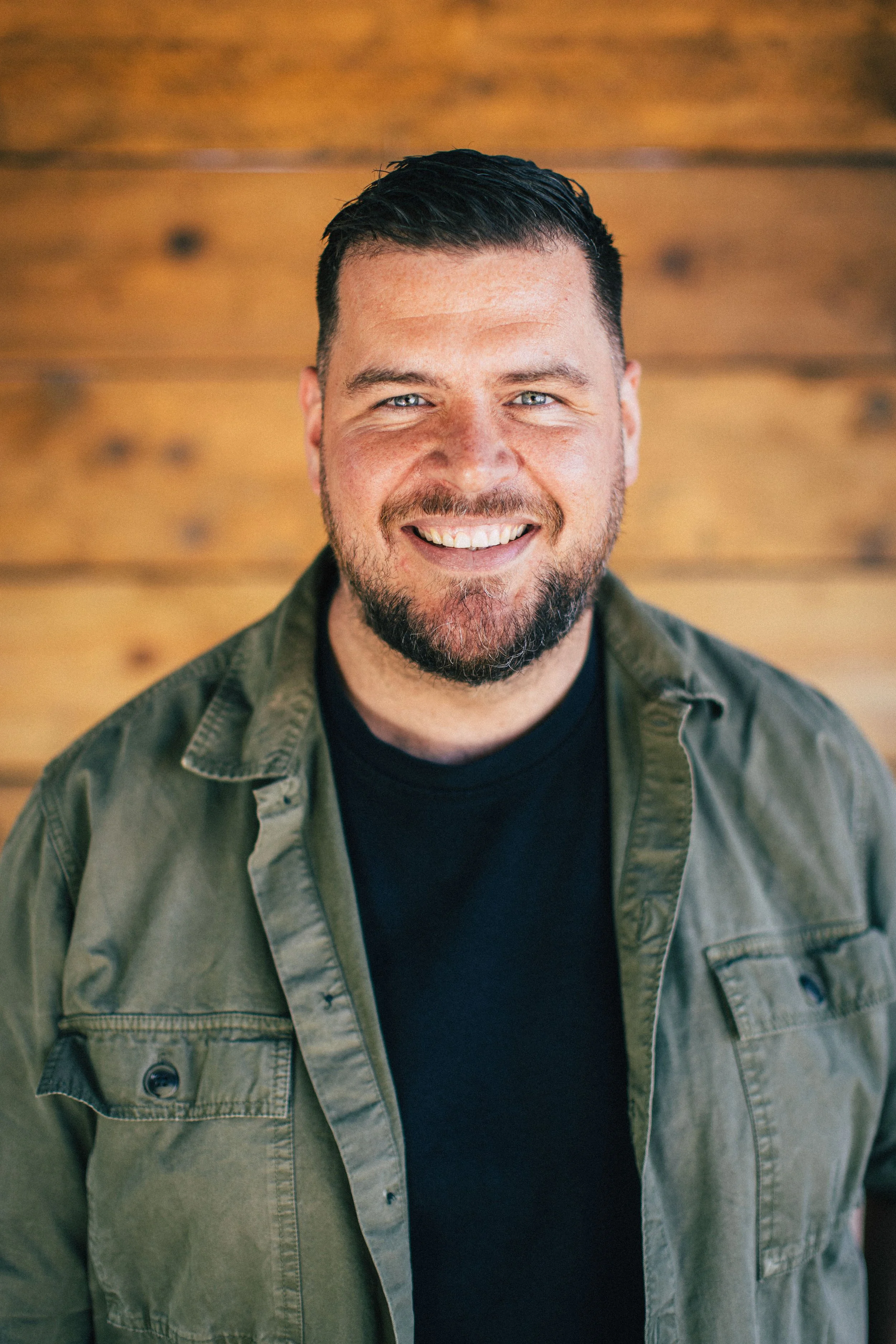 A smiling man with short dark hair and a beard, wearing a black t-shirt and an olive green jacket, standing in front of a wooden background.