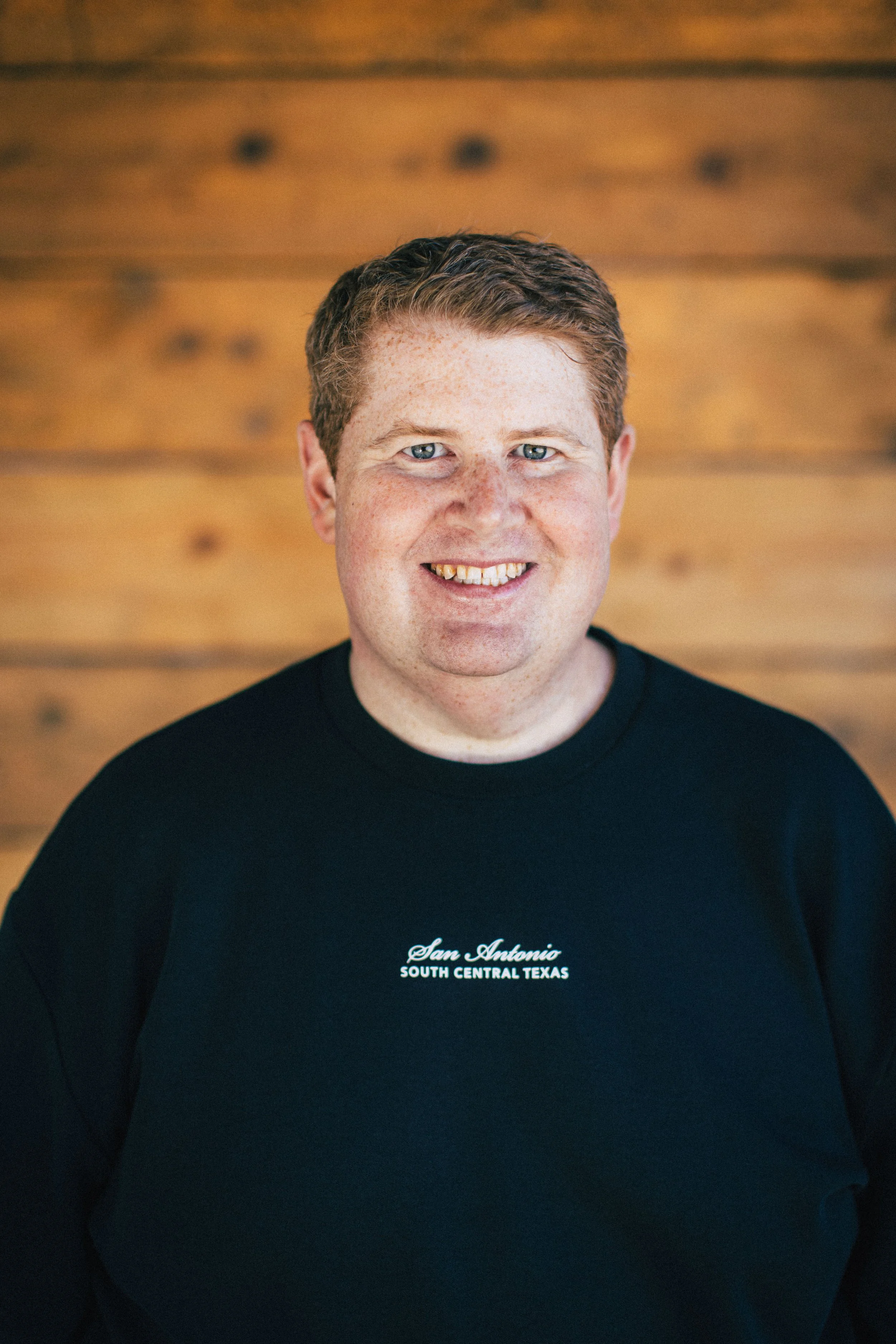 A man with red hair and freckles smiling at the camera, wearing a black shirt with 'San Antonio South Central Texas' embroidered on it, standing in front of a wooden wall.