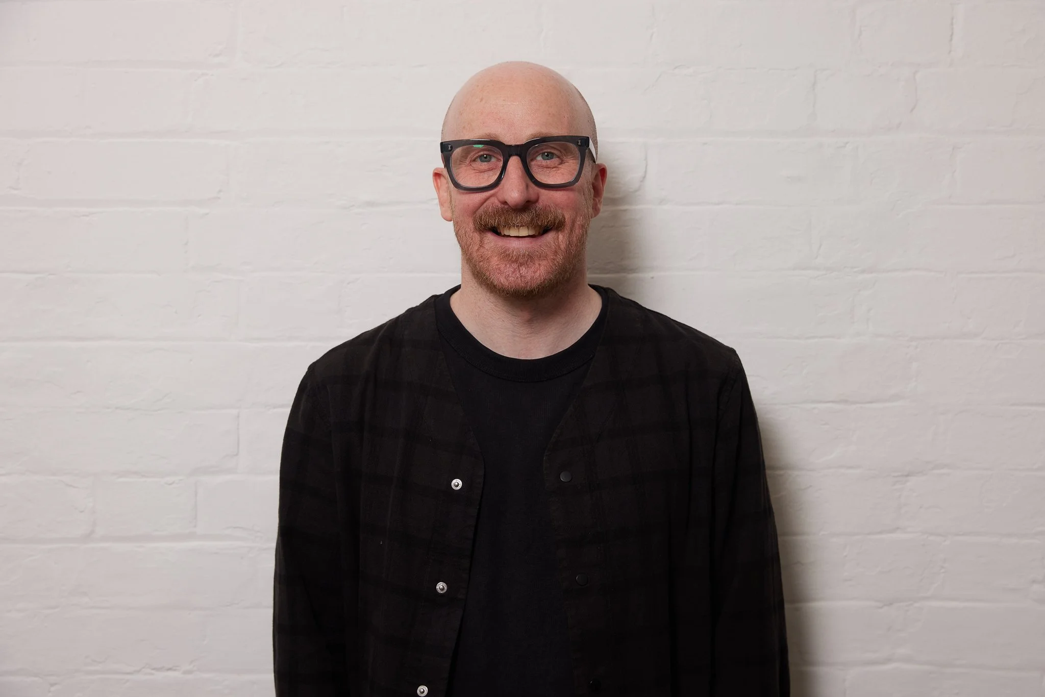 A smiling man with glasses and a beard, wearing a black T-shirt with a small yellow logo, standing in front of a wooden background.