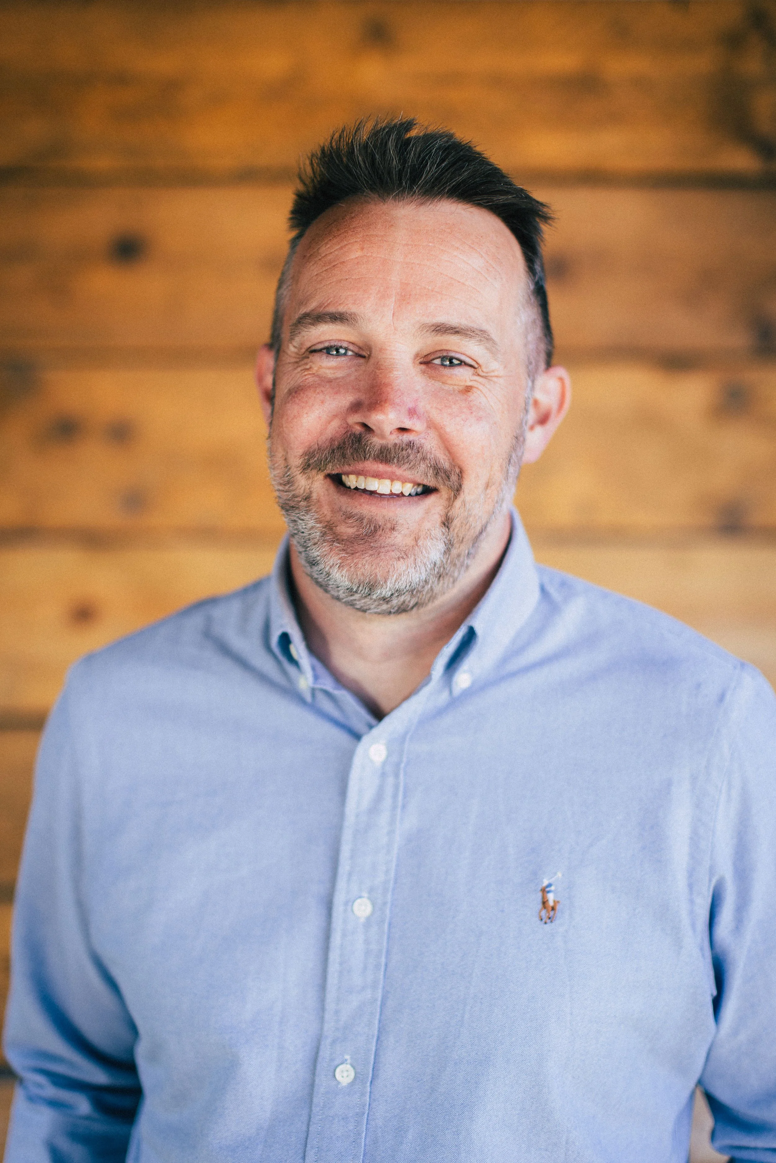 A smiling man with short dark hair, a beard, and blue eyes, wearing a light blue button-up shirt with a small horse logo, standing in front of a wooden wall.