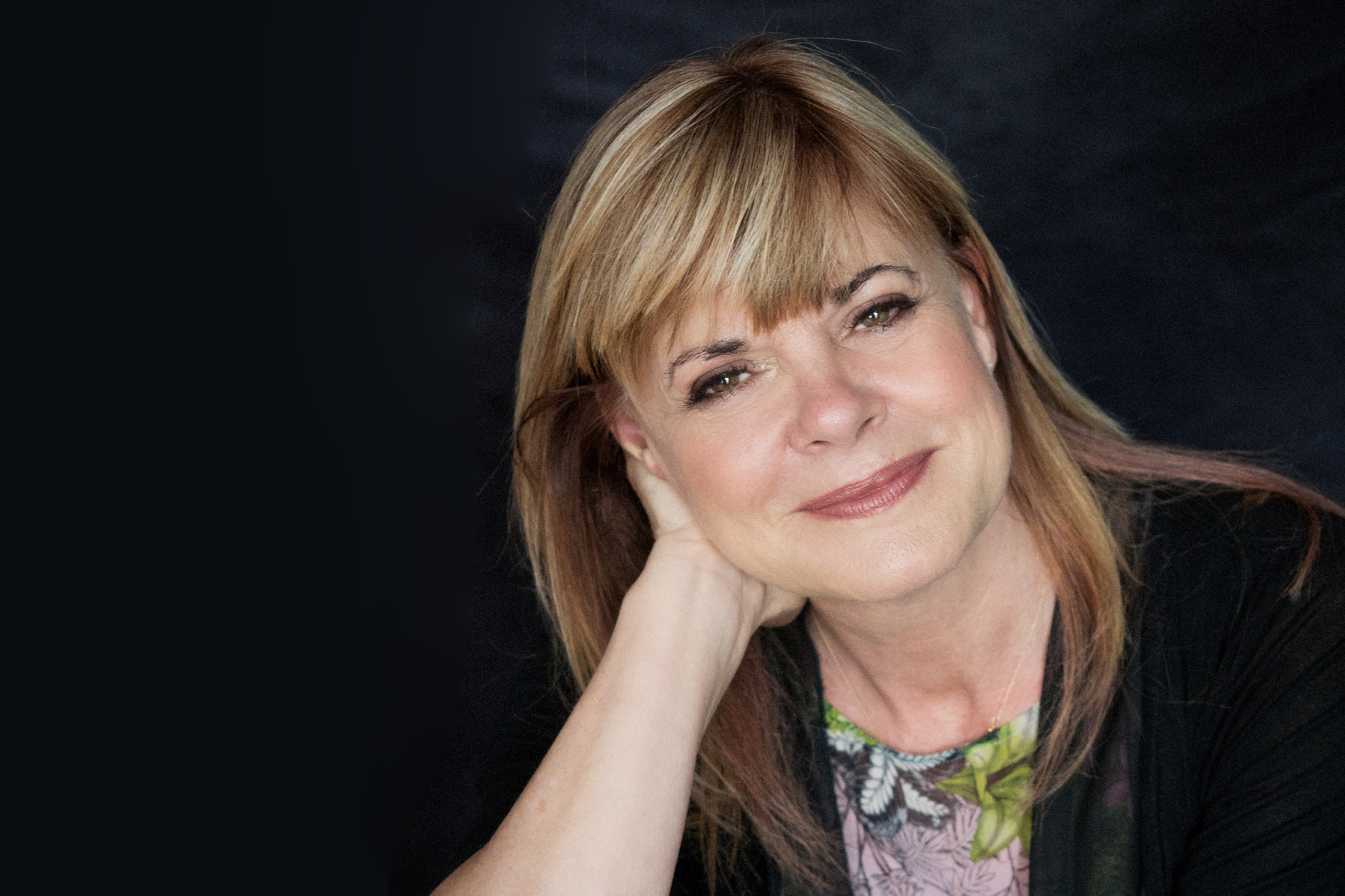 A woman with blonde hair, wearing a floral top and a black jacket, resting her hand on her cheek and smiling softly against a black background.