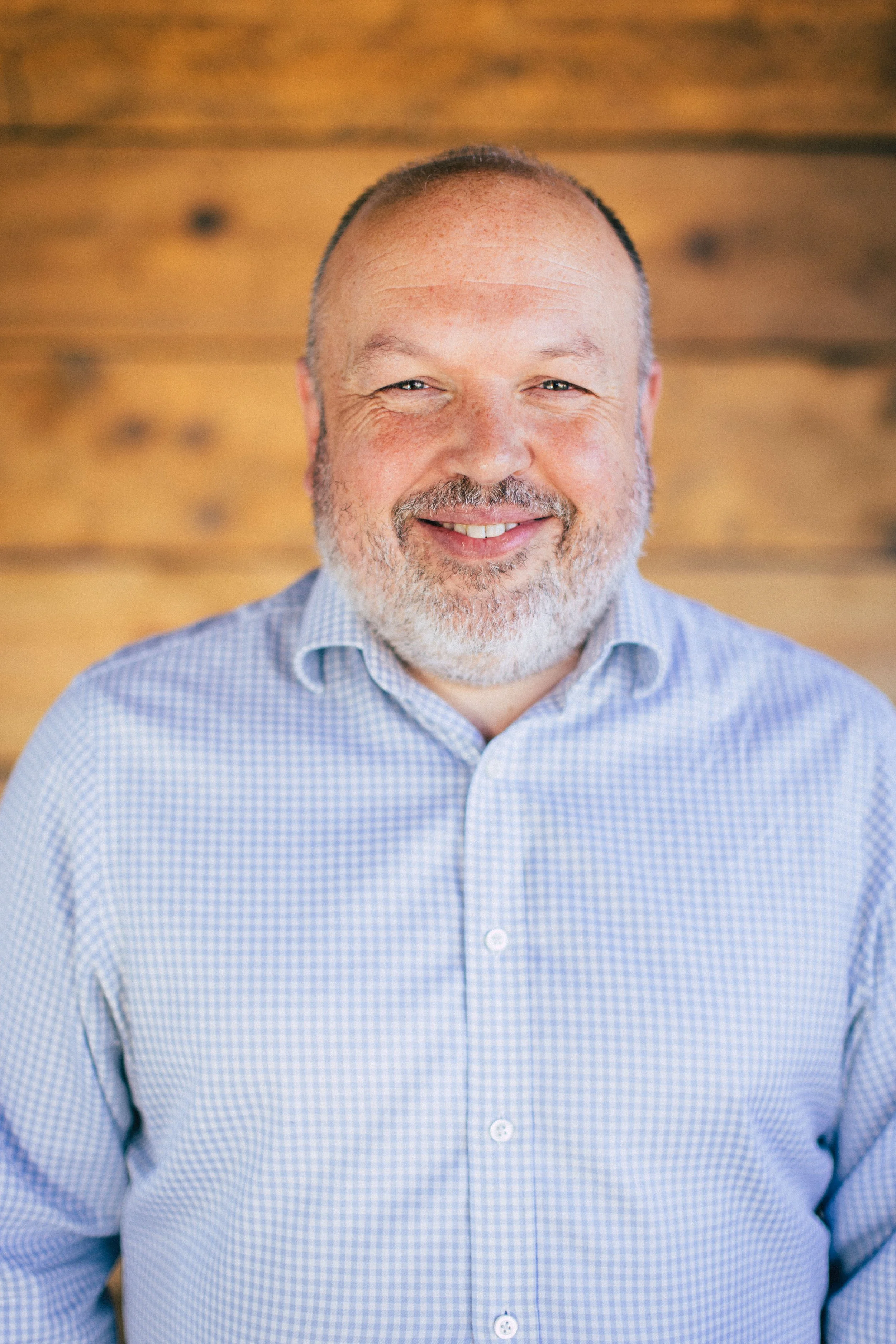 A smiling man with a beard and short hair, wearing a blue checkered shirt, in front of a wooden background.