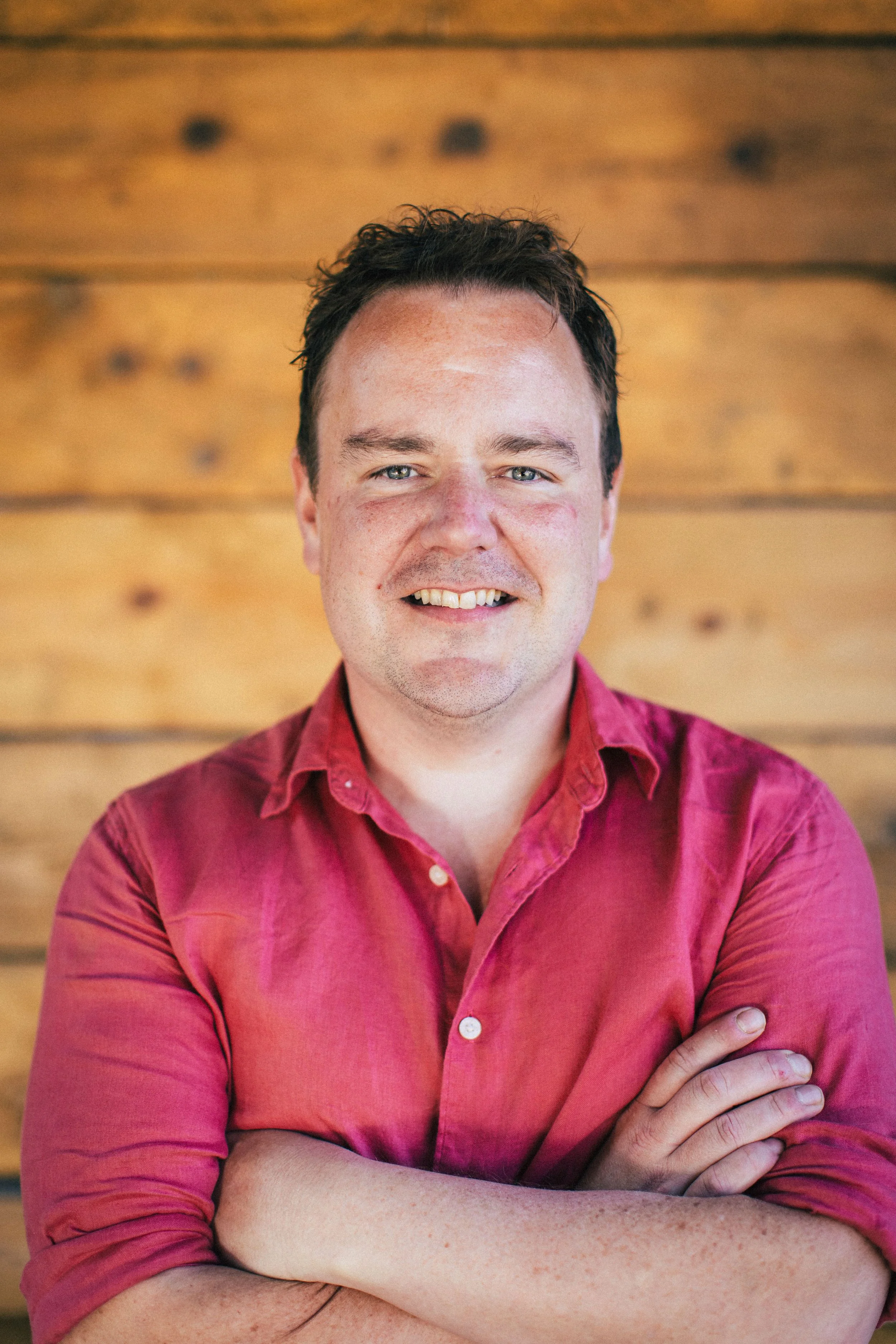 Portrait of a smiling man with arms crossed, wearing a red button-up shirt, standing in front of a wooden background.