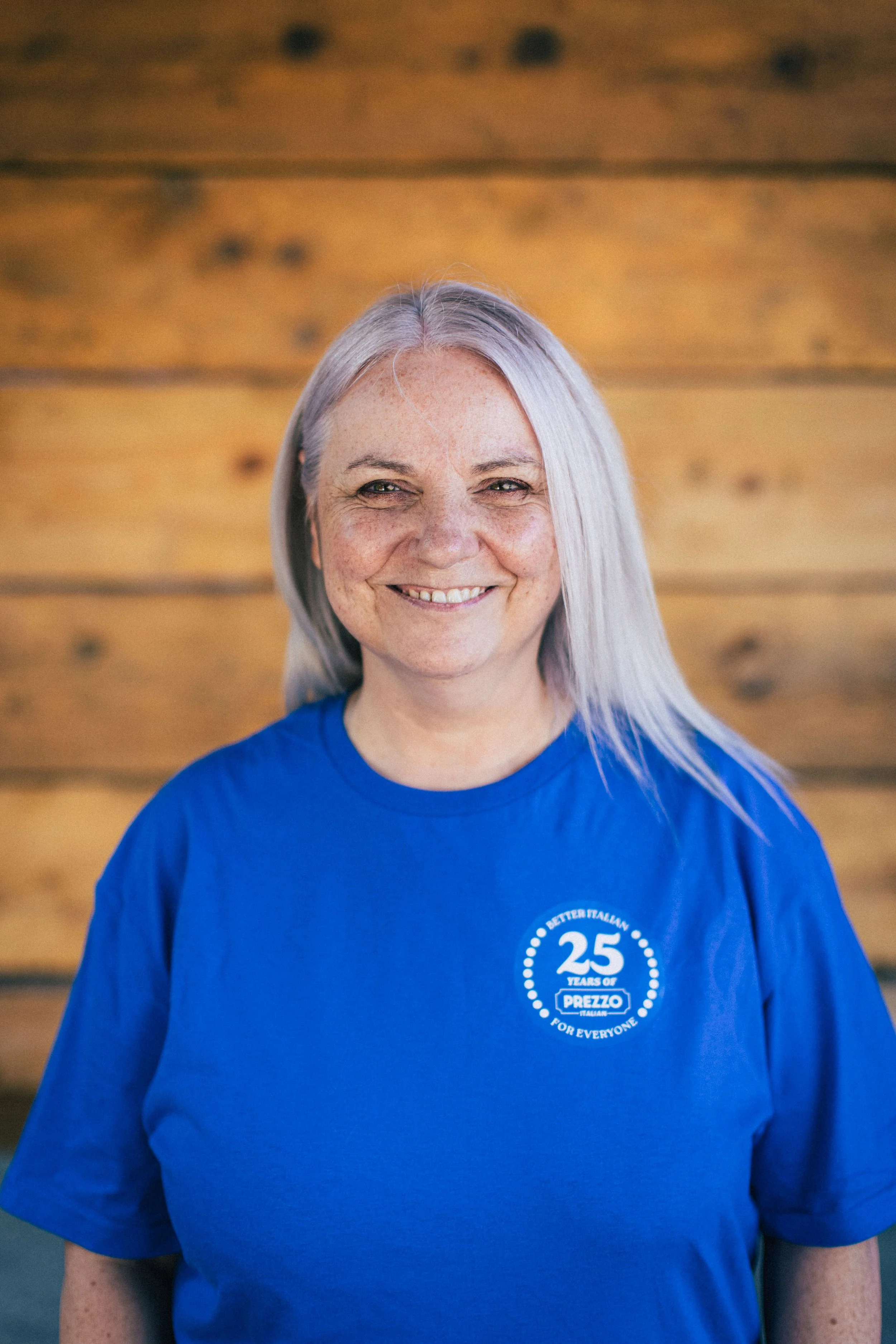 A smiling woman with long gray hair wearing a blue T-shirt in front of a wooden background.