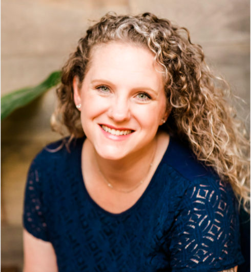 Portrait of a smiling woman with curly blonde hair wearing a navy blue lace top, sitting indoors near a wooden wall and green plant.
