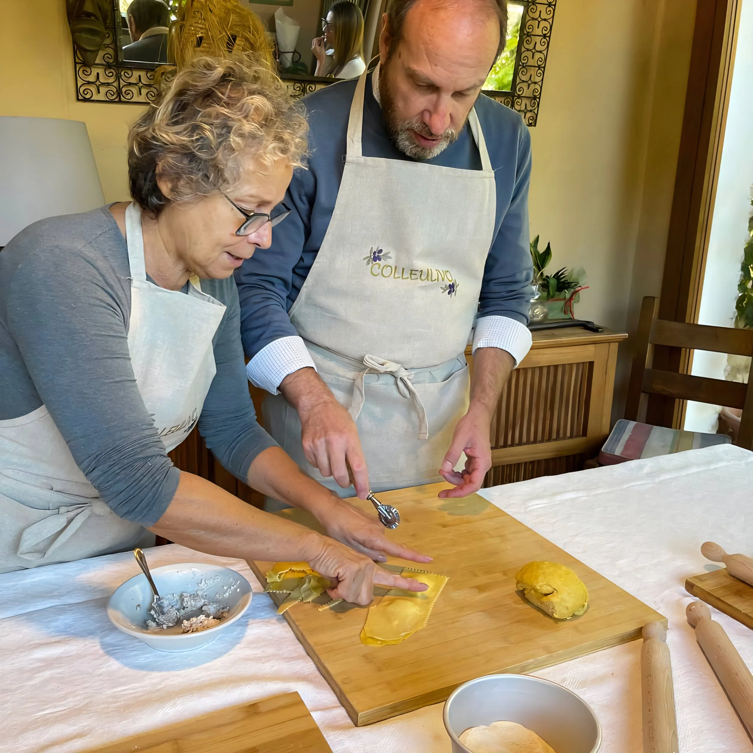 An elderly woman and a middle-aged man are cooking together in a kitchen, both wearing aprons. The woman is helping to cut a piece of pasta dough, while the man holds a pasta cutter. There are bowls, rolling pins, and dough on the wooden table.