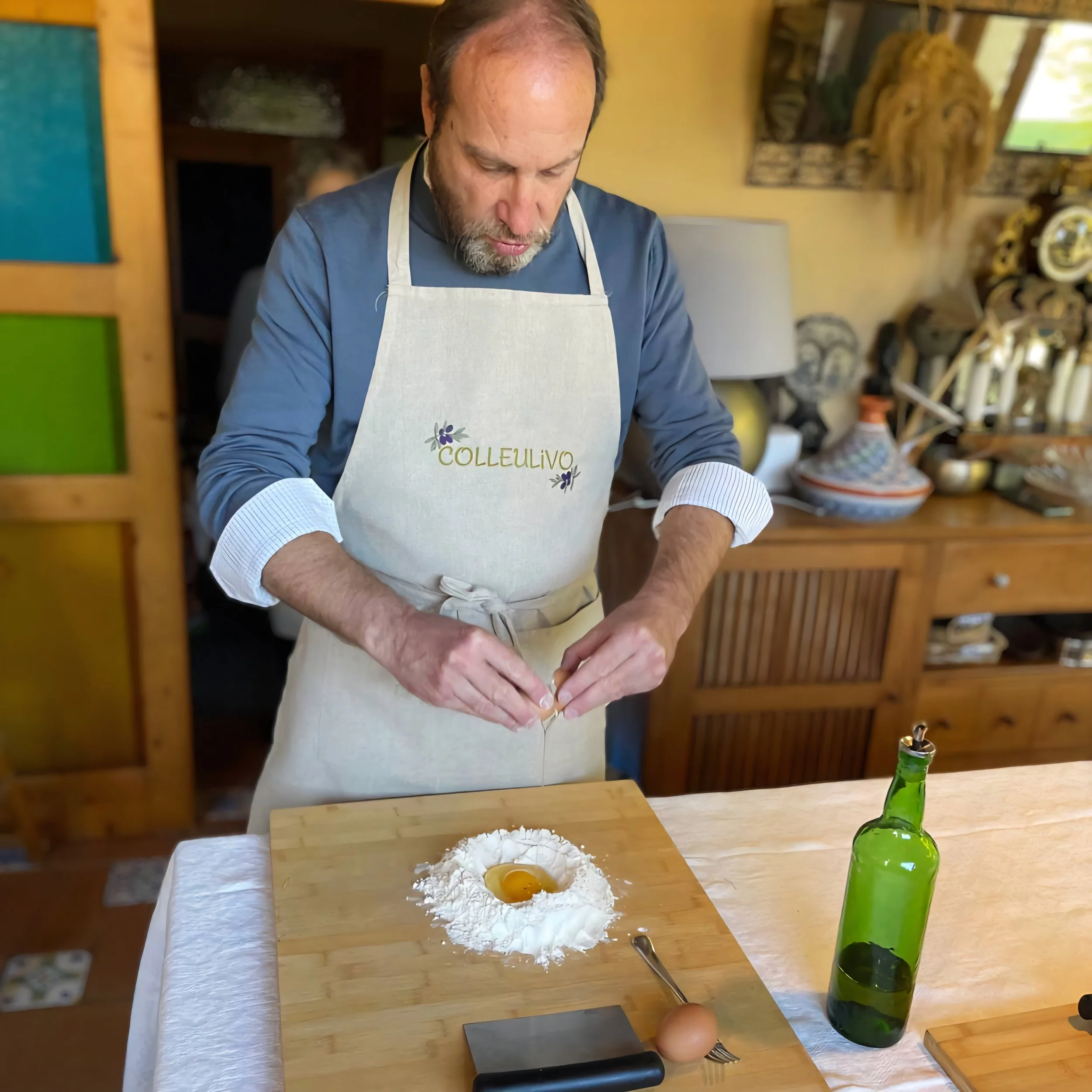 A man wearing a white apron with the word 'COLLEULIVO' stenciled on it is cracking an egg into a mound of flour on a wooden surface, preparing to cook or bake.