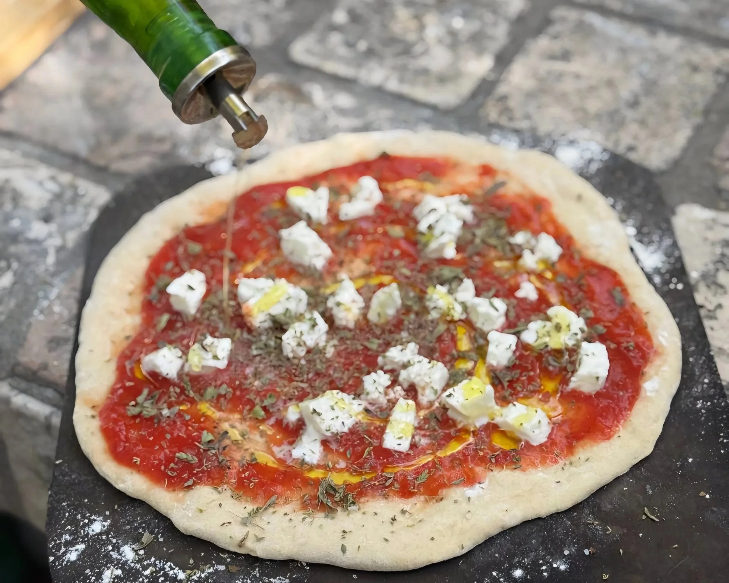 A pizza with tomato sauce, cheese, and herbs on a pizza stone. Olive oil is being drizzled on top.