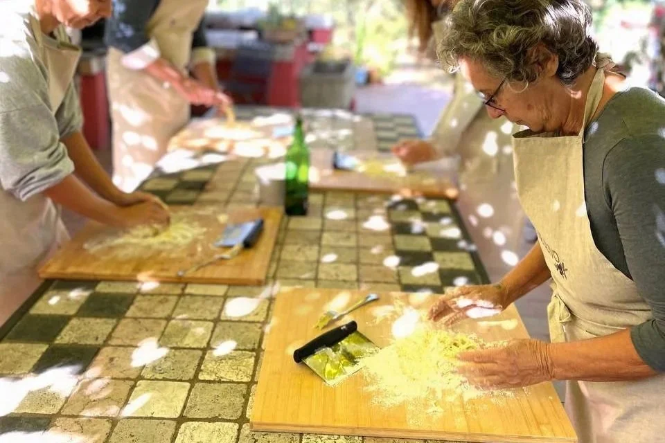 Several people participating in a cooking class, kneading dough on wooden cutting boards in an outdoor setting with dappled sunlight.