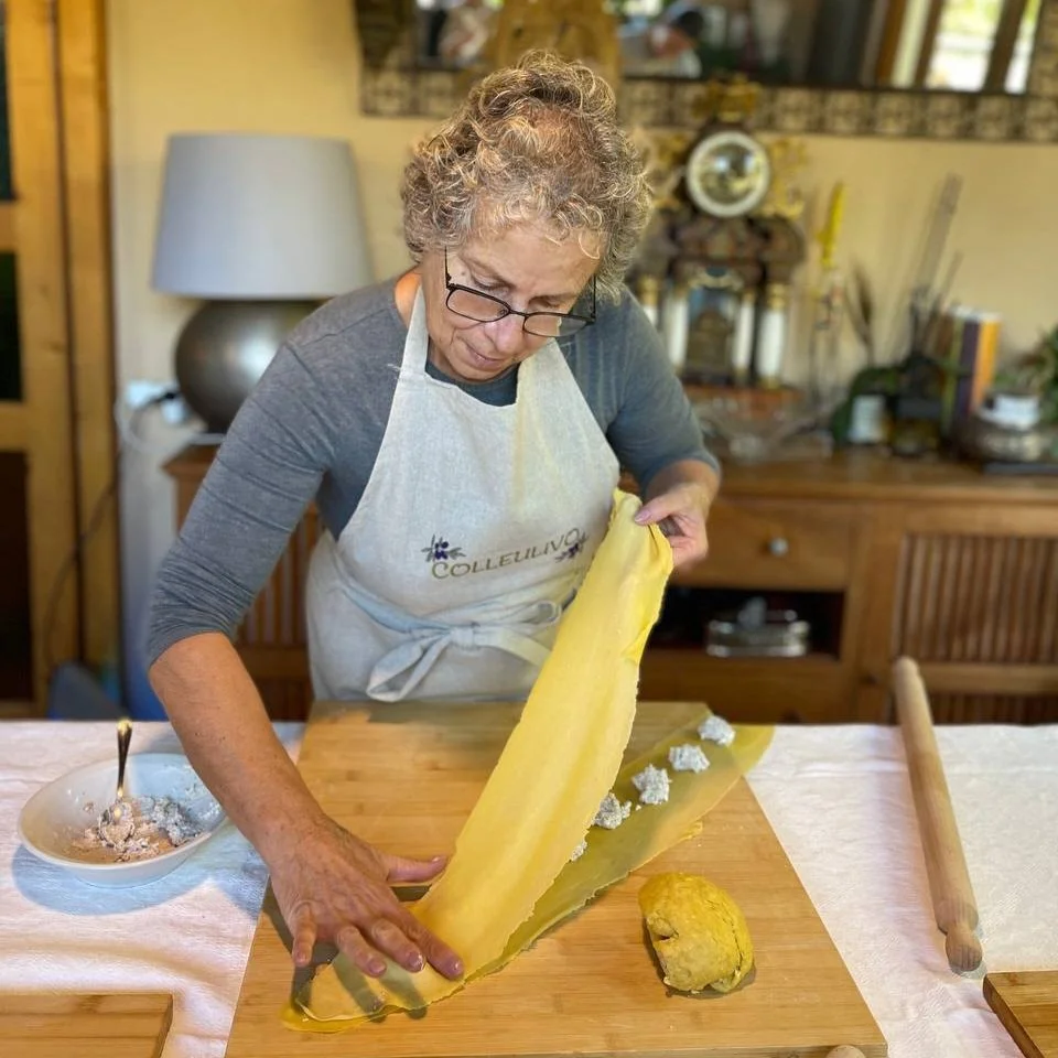 A woman, wearing glasses and a beige apron with the word 'COLLEULIVO' on it, is folding a large sheet of yellow dough or pastry on a wooden table.