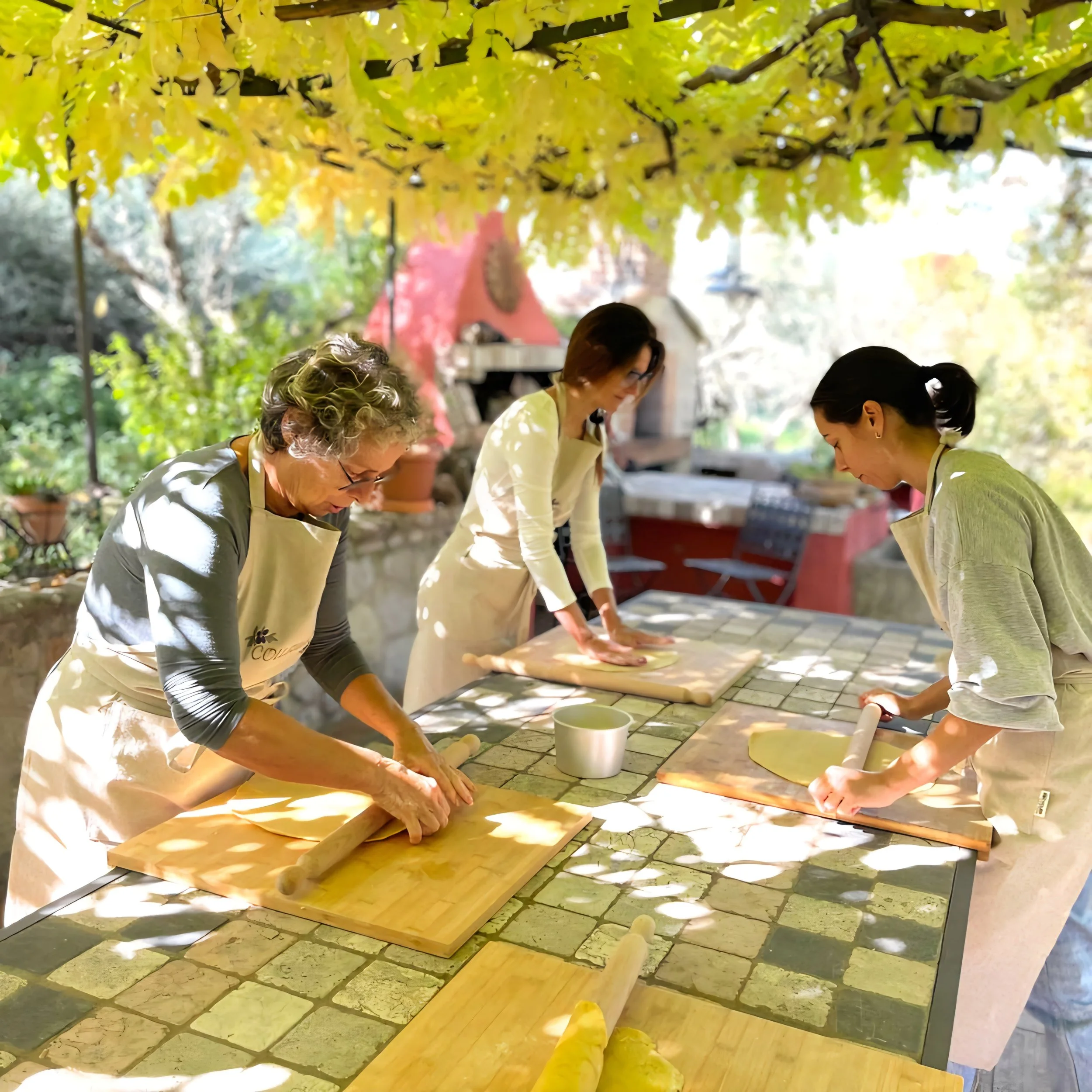 Three women baking dough outdoors under a leafy canopy on a sunny day.