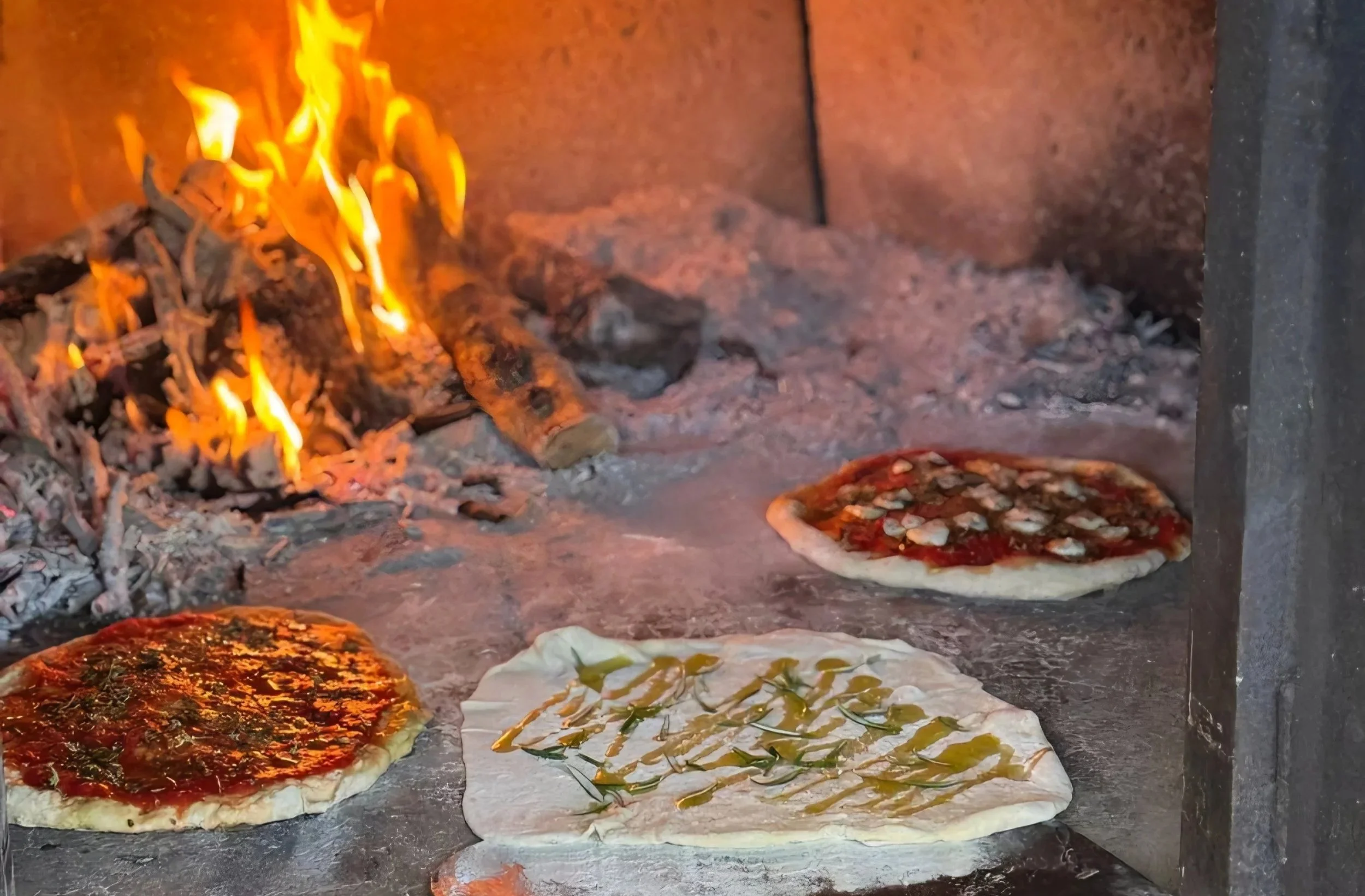 Three uncooked personal pizzas in a wood-fired oven with visible flames and burning logs in the background. The pizzas are topped with sauce, cheese, and various toppings, with one having a drizzle of green olive oil.