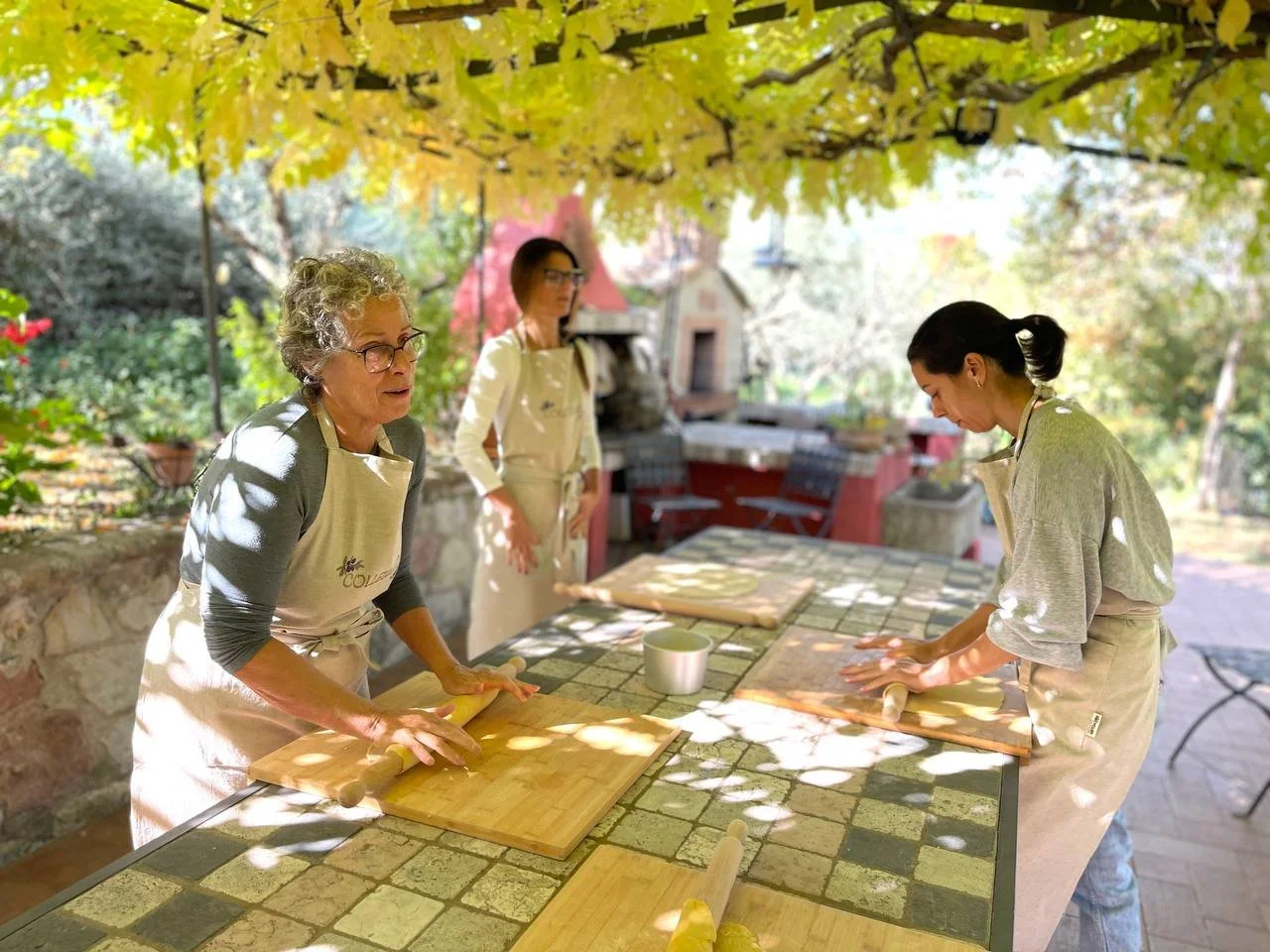 Three women cooking outdoors on a sunny day. Two women are rolling dough on wooden boards, and a third woman is watching. They are under a leafy canopy, with a rustic stone wall and garden in the background.