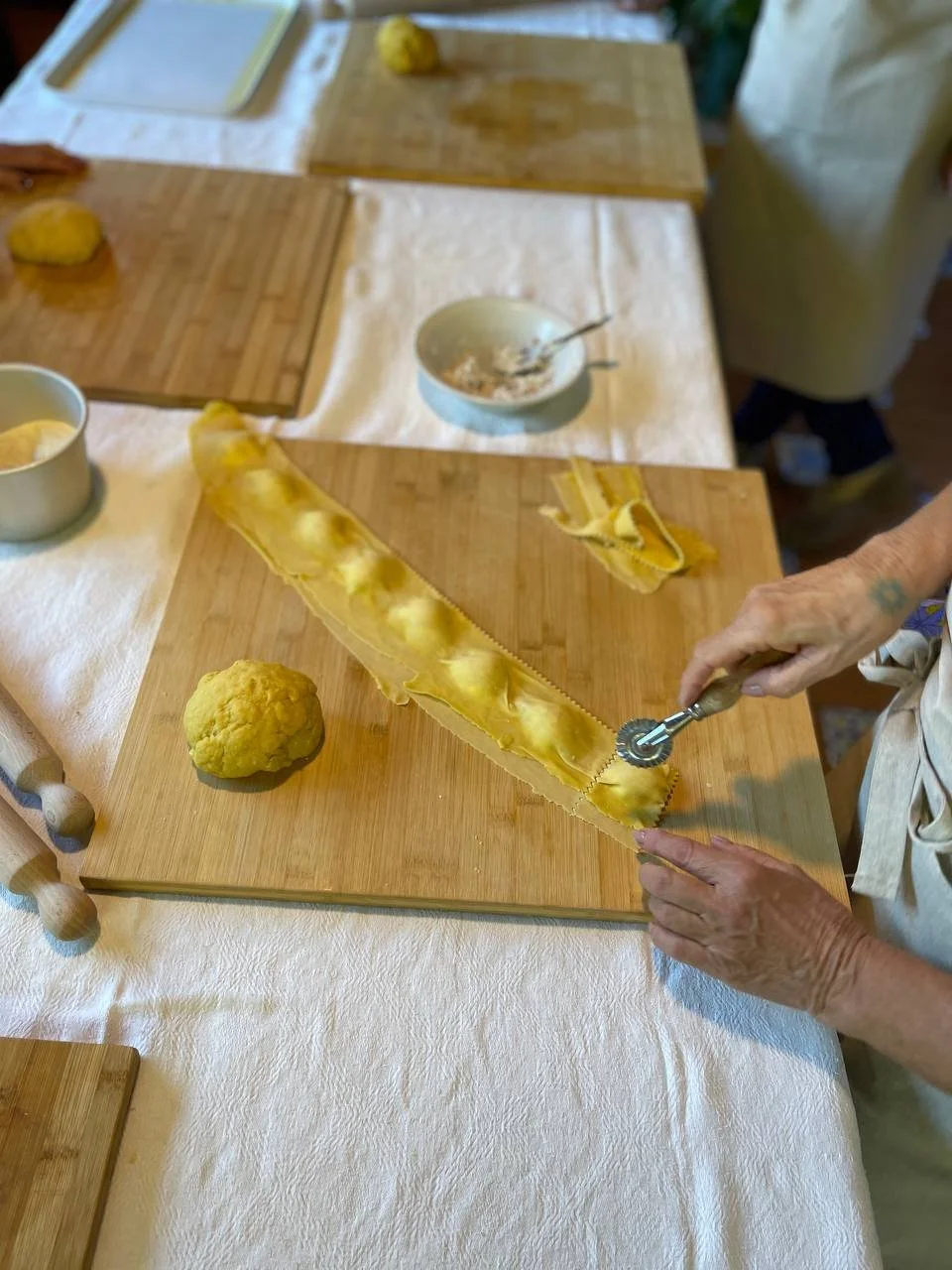 Person slicing a long, yellow pasta sheet with a scalloped-edged pasta cutter on a wooden cutting board in a kitchen. A ball of yellow dough and other kitchen tools are also visible on the table.