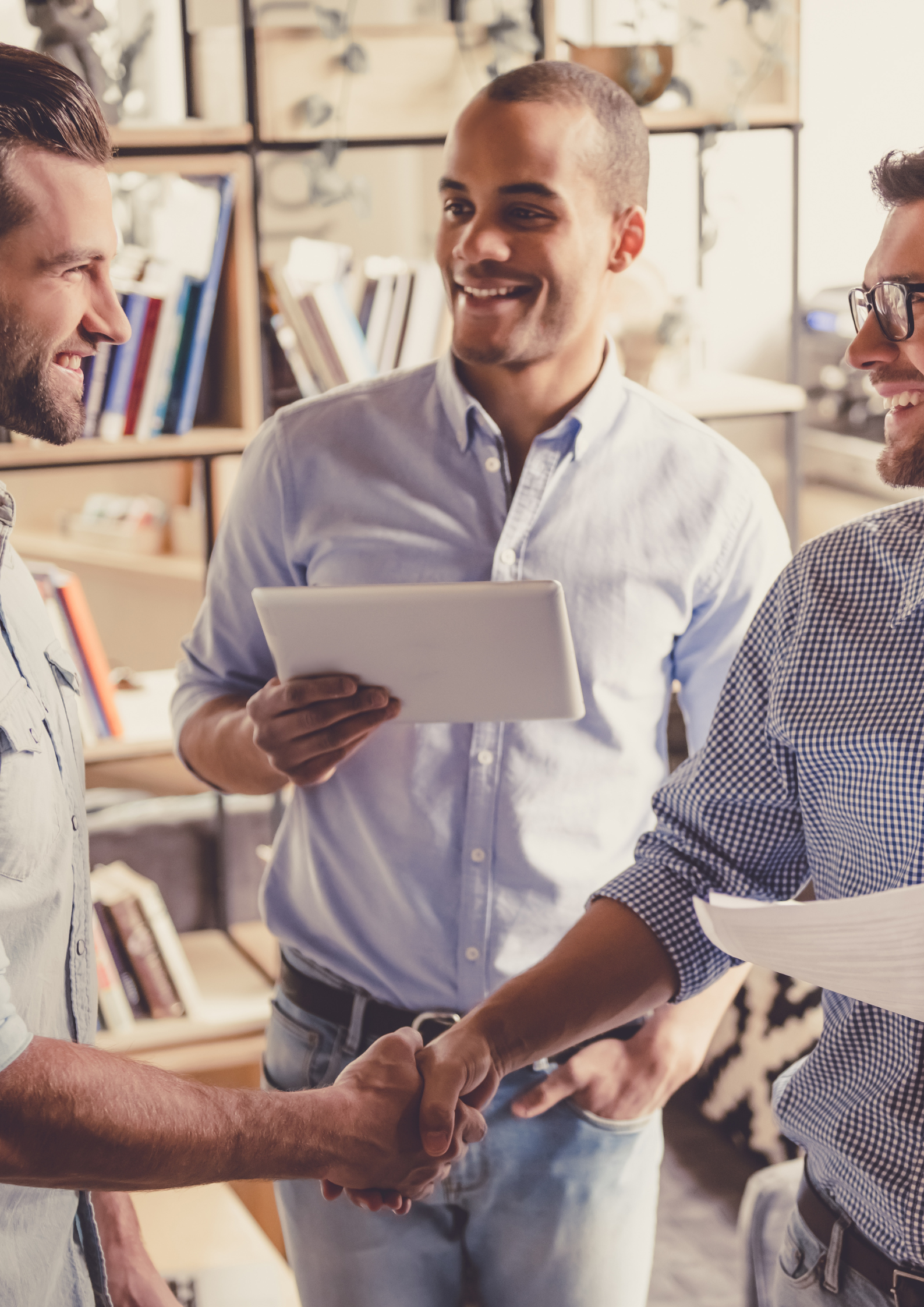 Three men smiling and shaking hands in a professional setting, with one man holding a tablet and another holding some papers.
