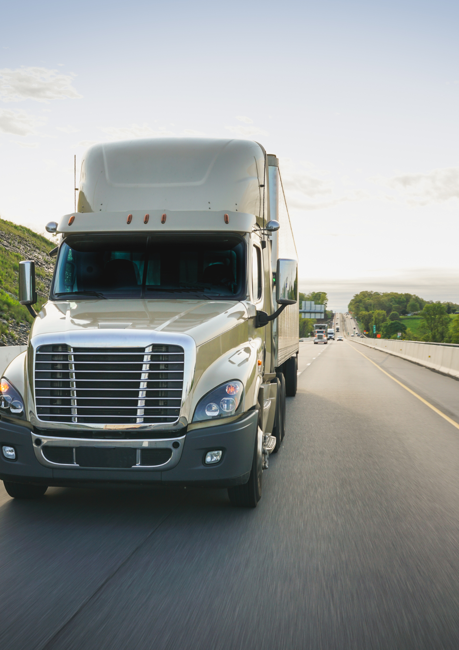 A beige semi-truck driving on a highway with green trees and hills in the background on a clear day.