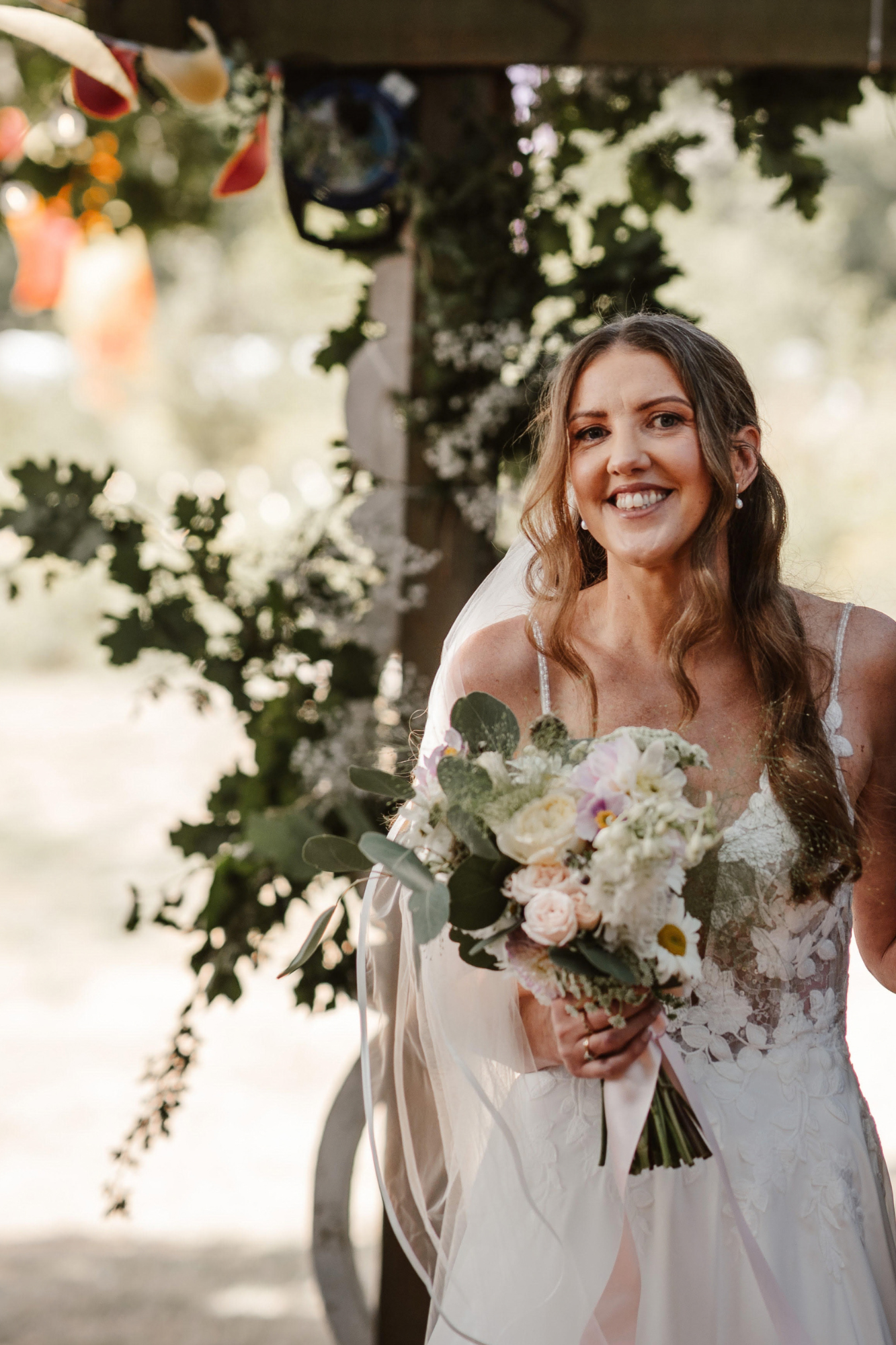 A bride holding a wild flower insired bouquet of white and pale pink roses and dahlias amoungst other flowers and foliage, smiling outdoors during her wedding, under a wooden arch decorated with greenery and white flowers.
