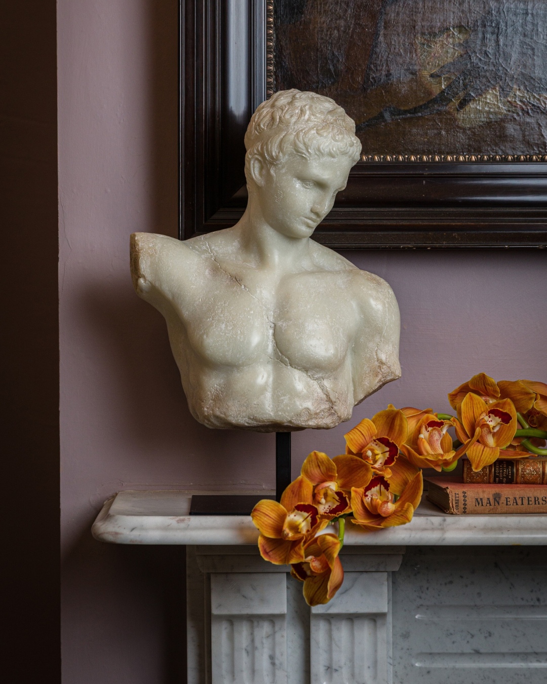 Classical bust of a man on a white marble mantel, styled with orange orchids draping down from antique books.