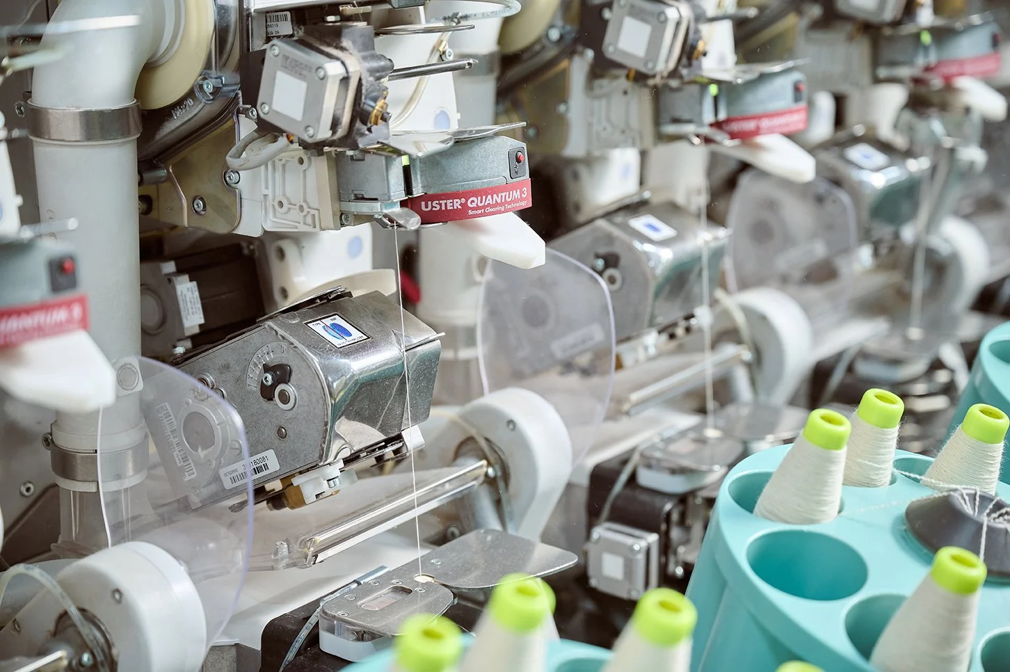 Close-up of a textile machine with spools of white thread and sewing components, labeled 'USTER QUANTUM 3'.