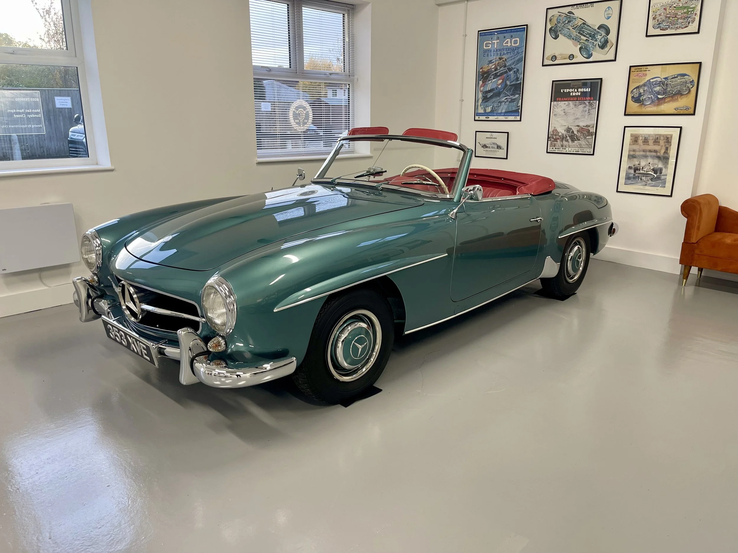 Vintage Mercedes-Benz convertible car with a teal body and red interior, displayed indoors near framed car posters.