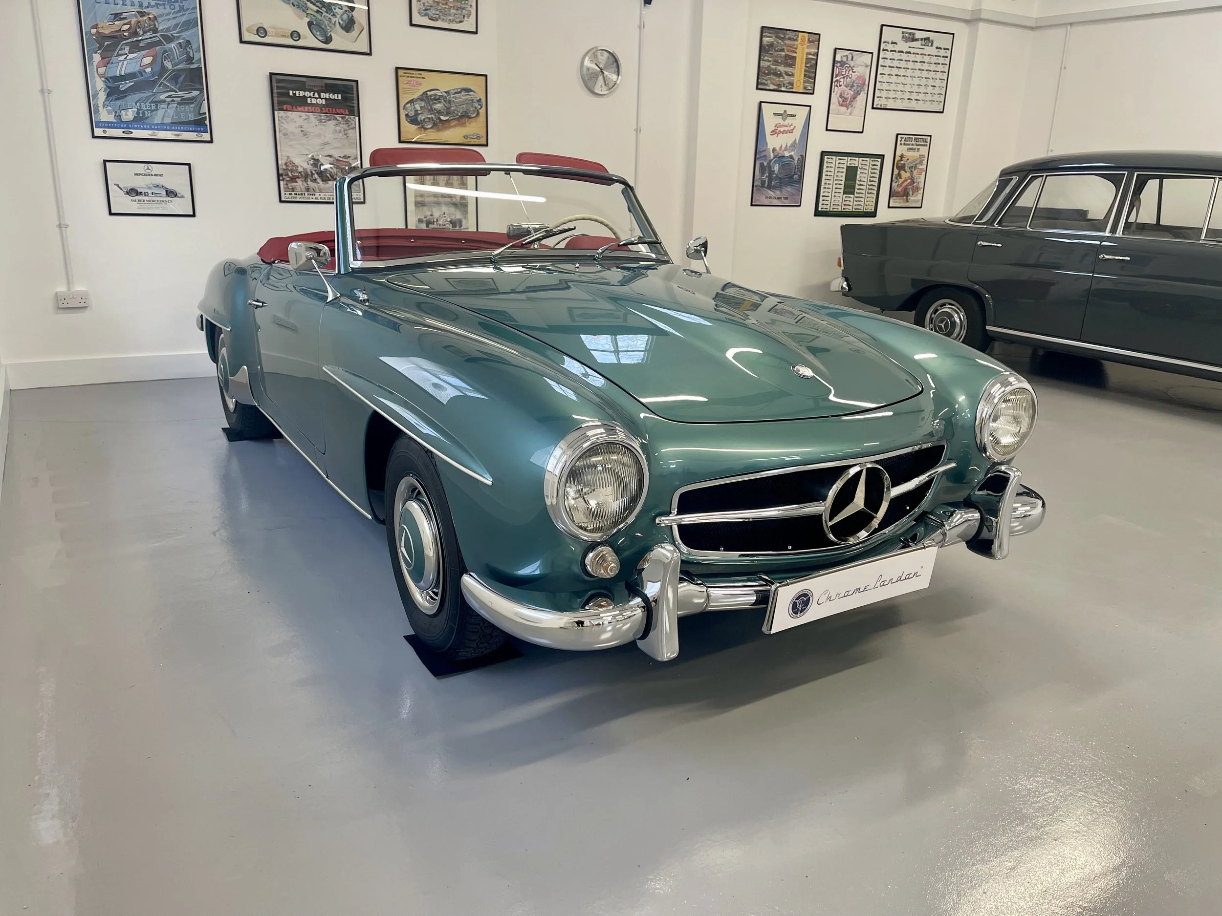 A vintage green Mercedes-Benz convertible car with a red interior parked inside a museum, with framed posters and car photographs on the white wall behind it.