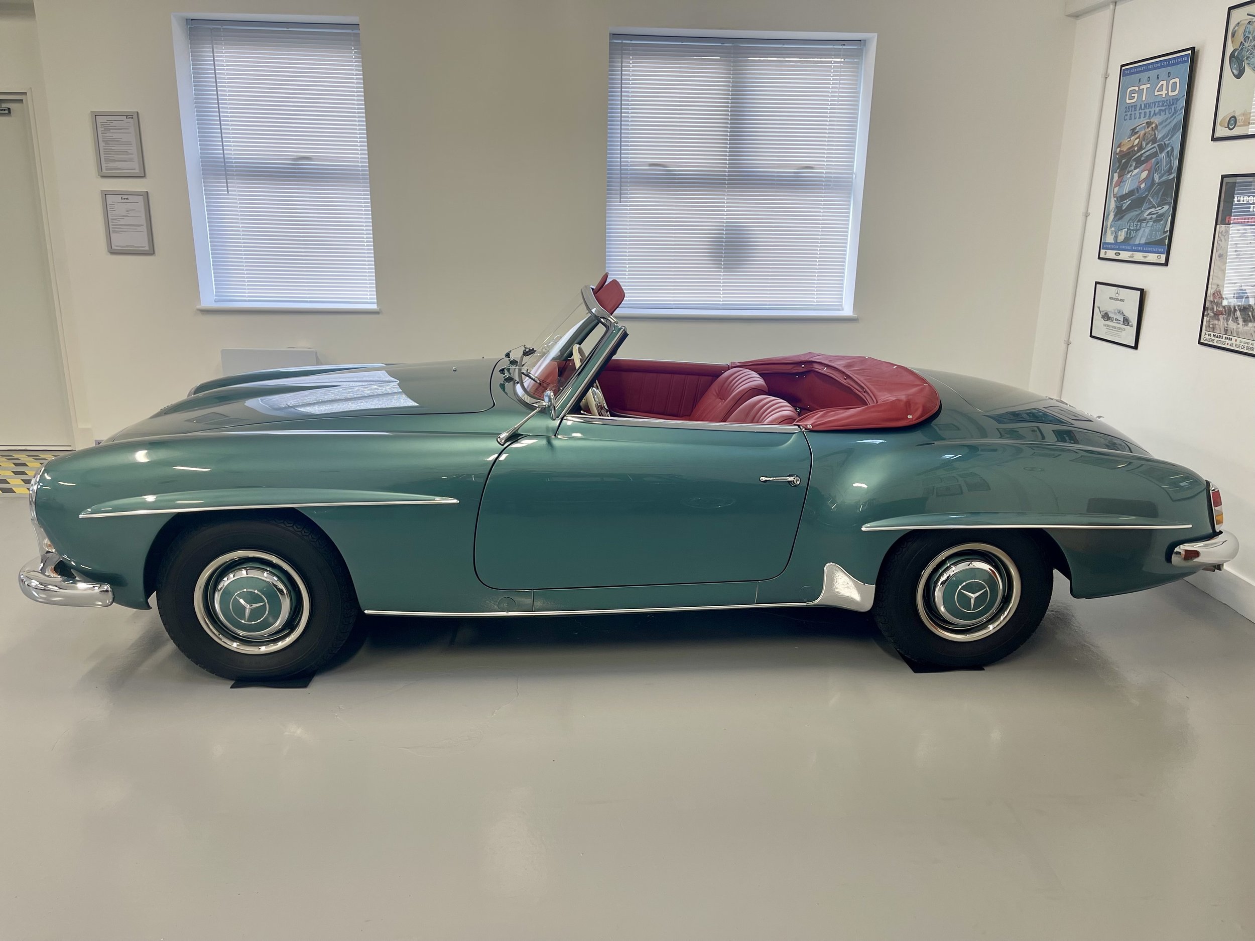 A vintage green Mercedes-Benz convertible with a red interior, displayed indoors, with framed vehicle posters on the walls.