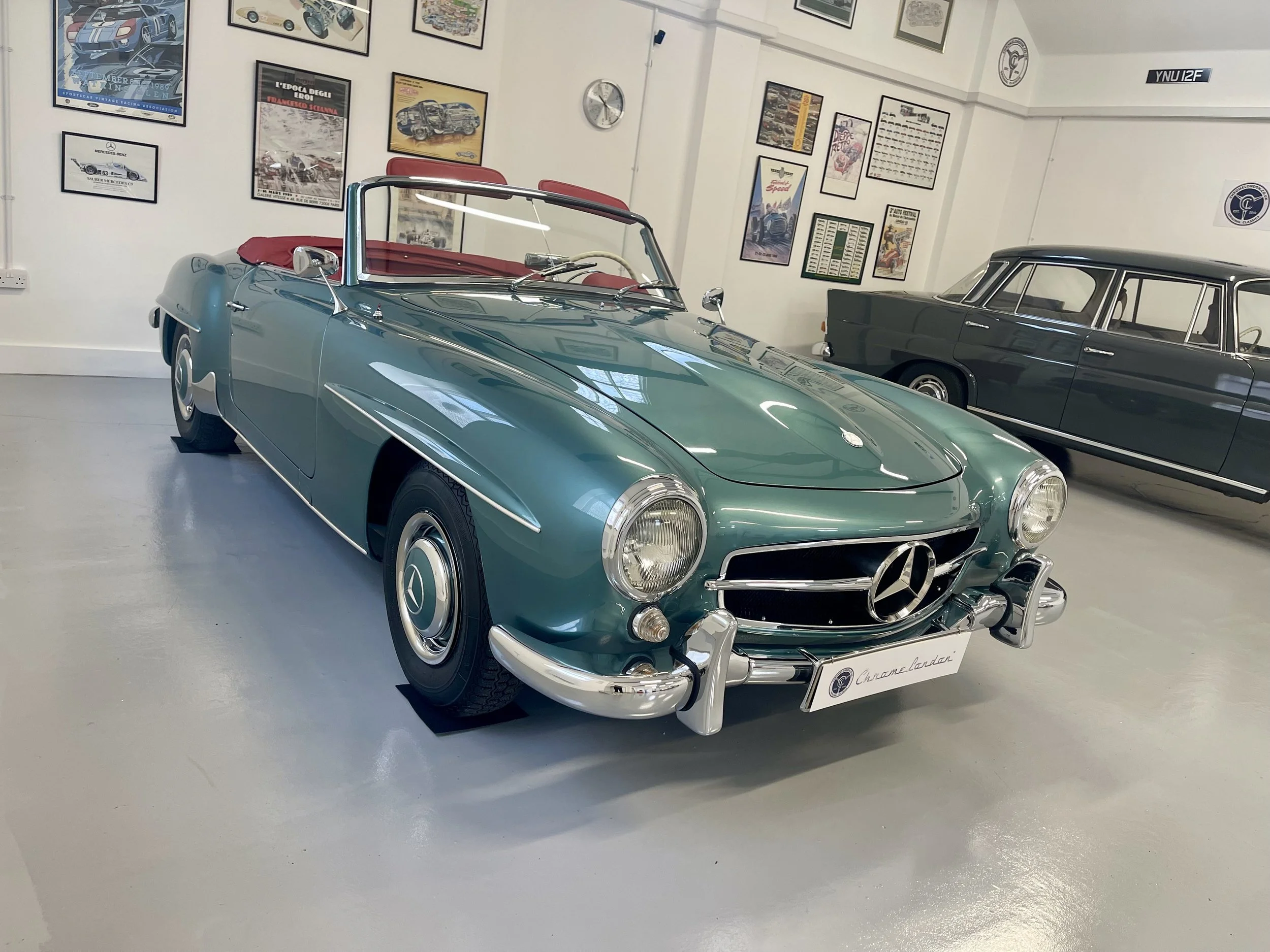 A vintage teal Mercedes-Benz convertible car with a white interior displayed in a museum, with framed car posters on the walls behind it.