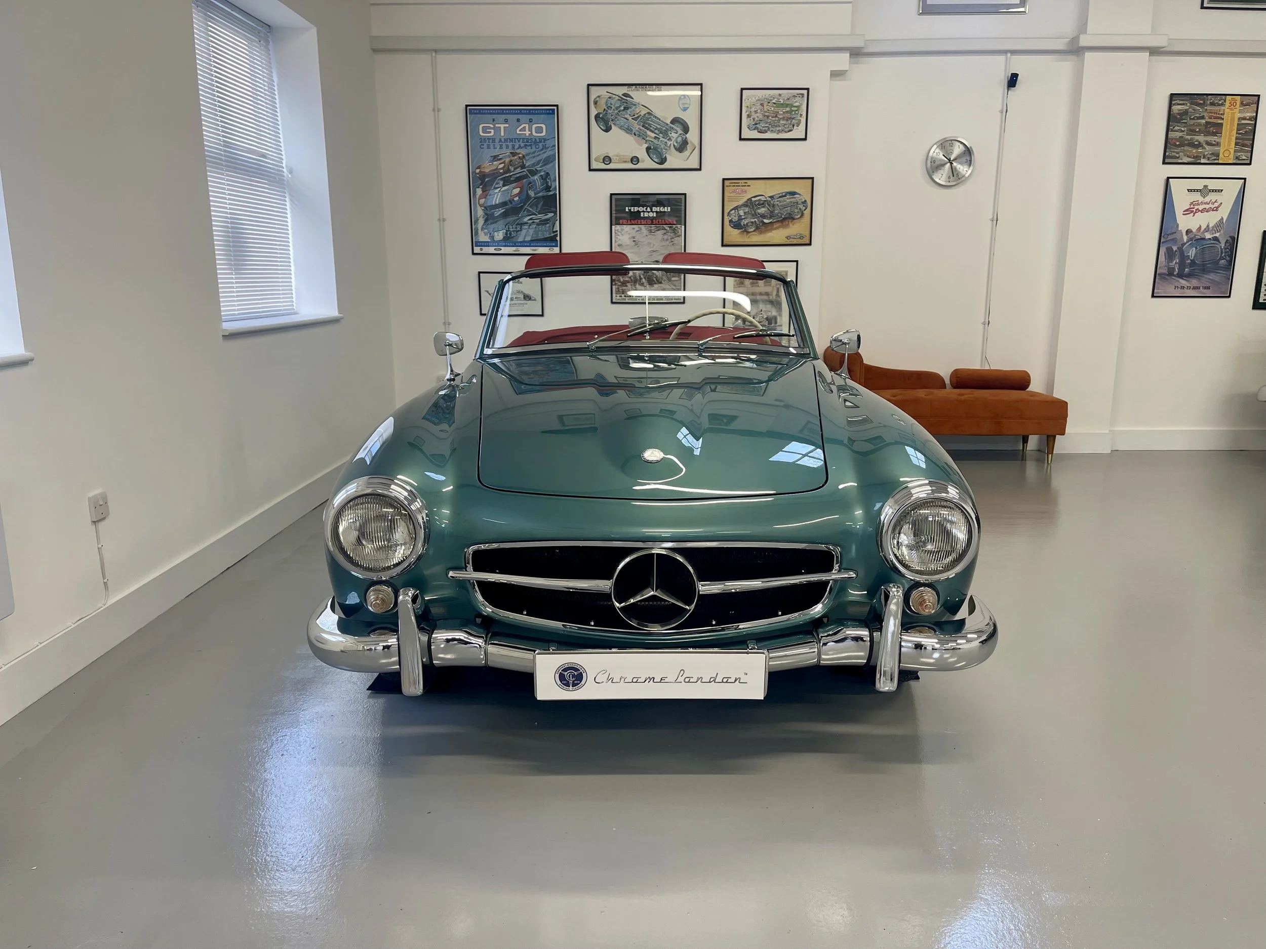 A vintage teal Mercedes-Benz convertible car with a red interior displayed indoors, surrounded by framed posters and pictures of cars on the walls.