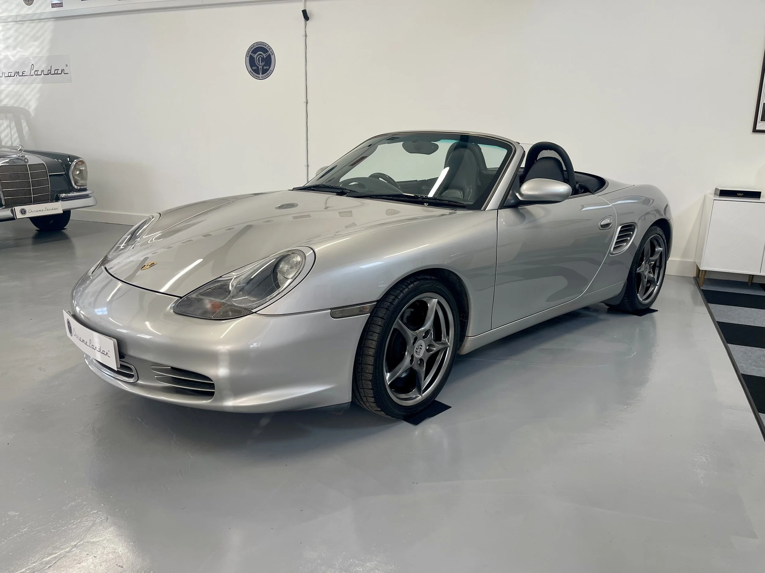 A silver Porsche Boxster convertible sports car on display in a showroom, with a classic Mercedes-Benz partially visible in the background.