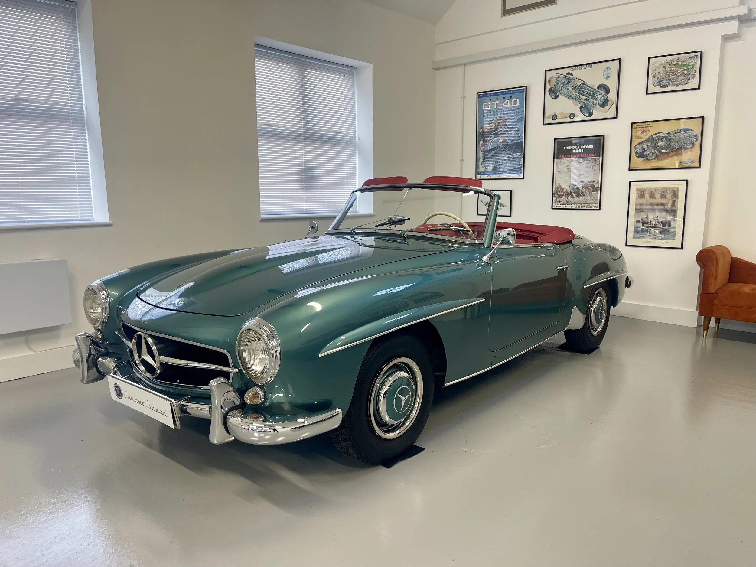Vintage green Mercedes-Benz convertible car with red interior parked indoors, framed automotive posters on the wall, and windows with blinds.