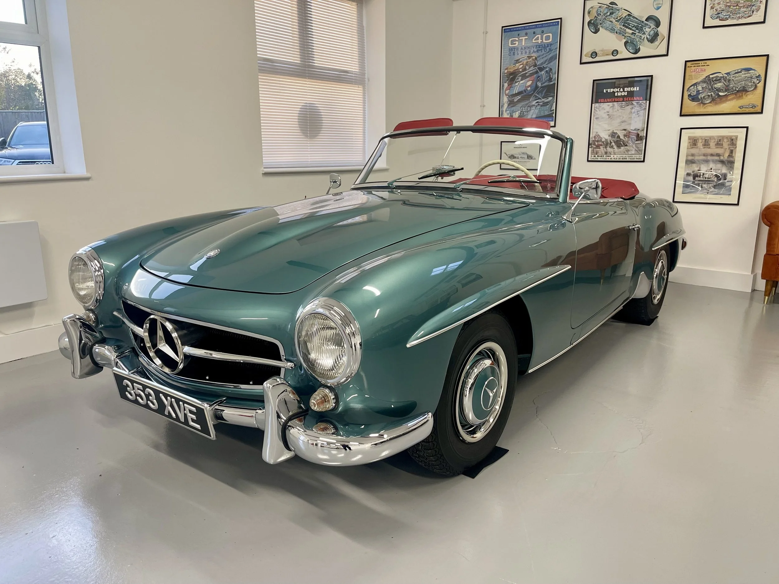 A vintage green Mercedes-Benz convertible car with red interior, parked indoors with framed posters of cars on the wall behind it.