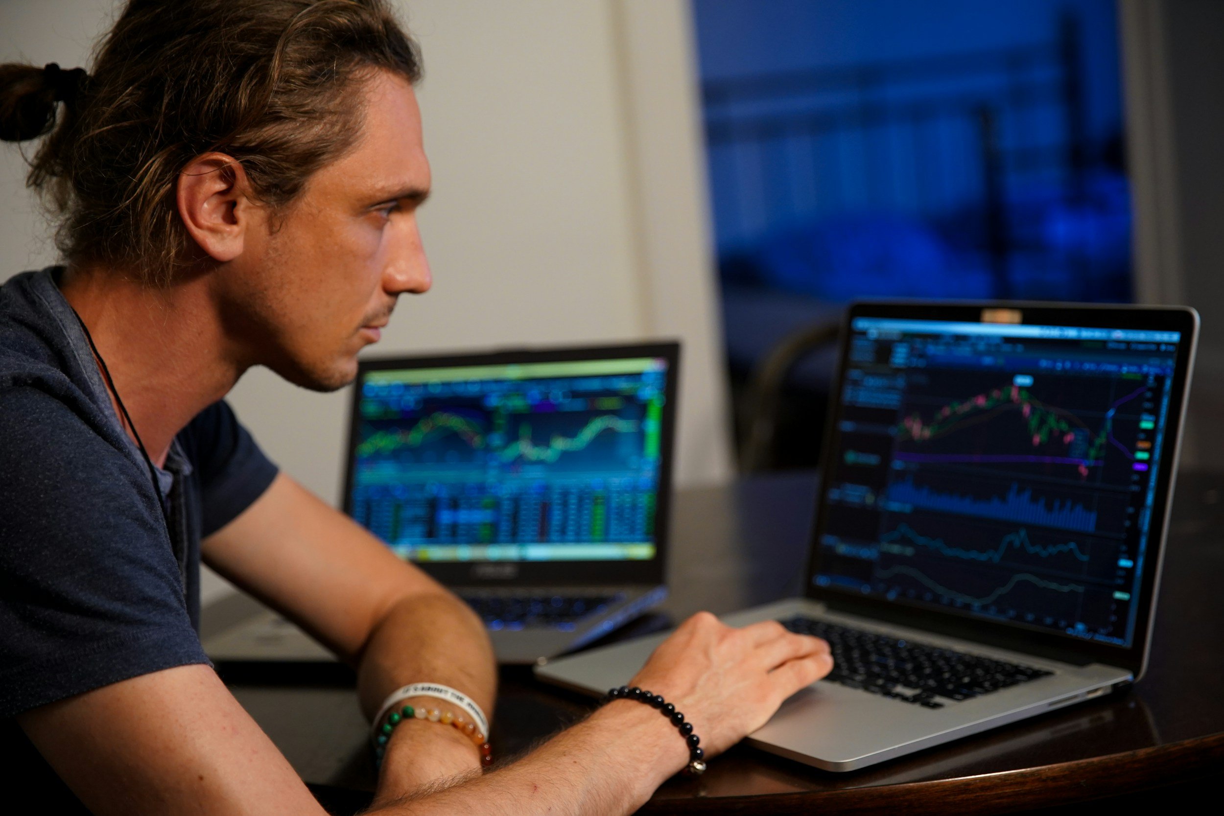 A young man with curly hair, wearing a dark t-shirt and beaded bracelets, is sitting at a desk working on a laptop with financial charts on the screen, in a dimly lit room during evening.