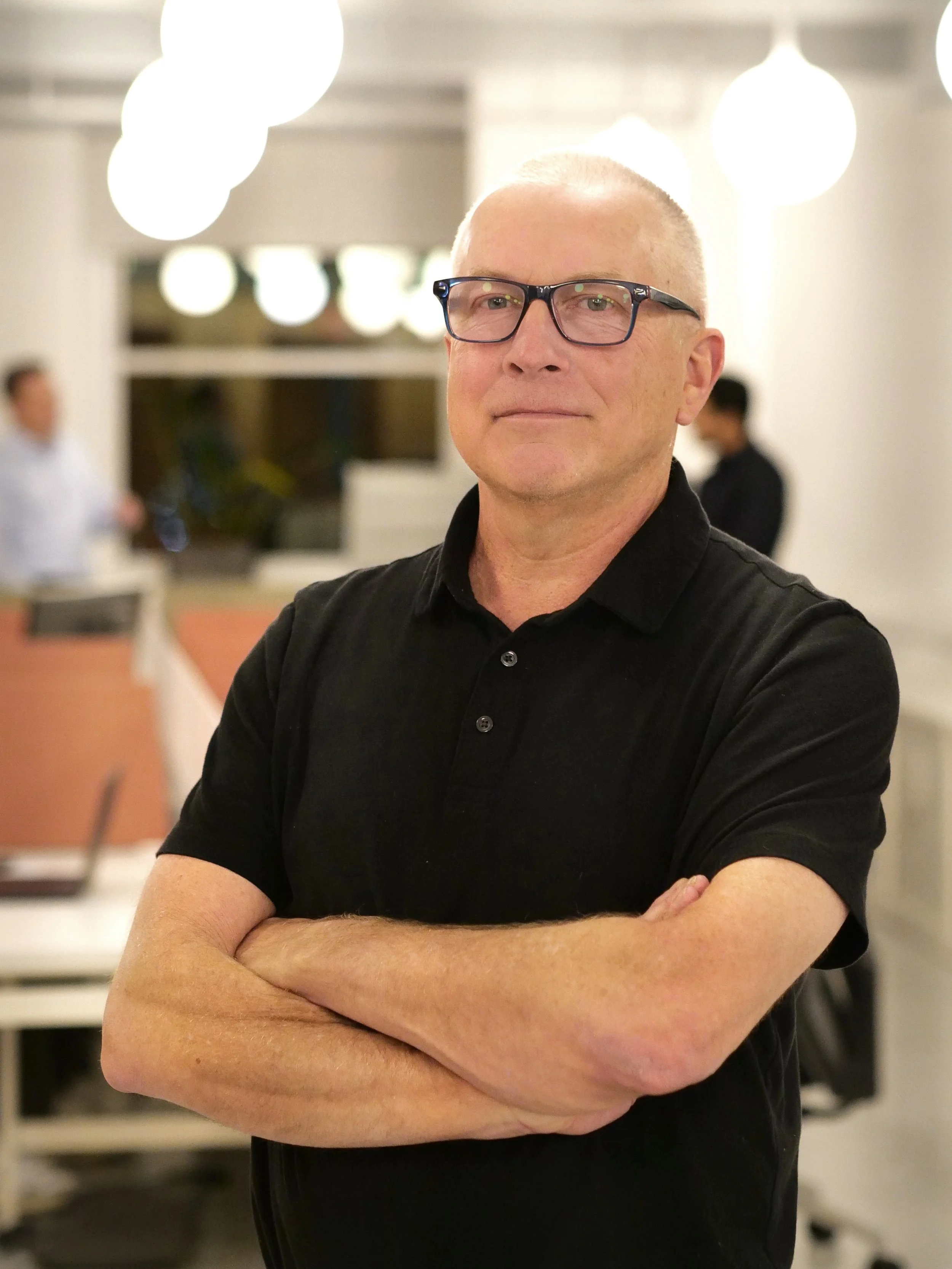 A man with short gray hair and glasses wearing a black blazer and a blue checkered shirt, smiling in a bright modern office building.