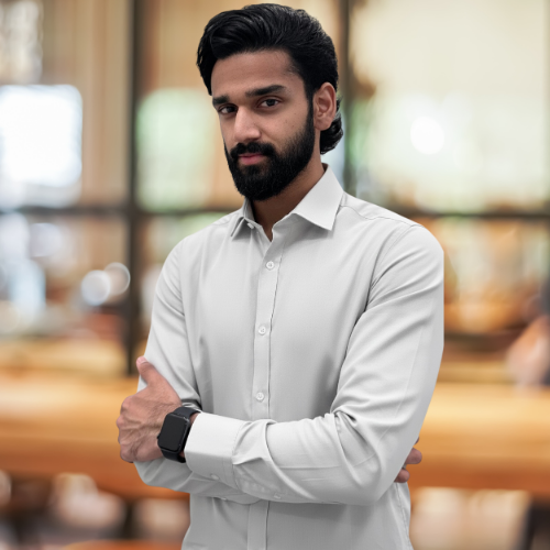 A man with dark hair, a beard, and a mustache wearing a beige suit, white shirt, and black tie, standing in a modern office building with large windows and blurred background.
