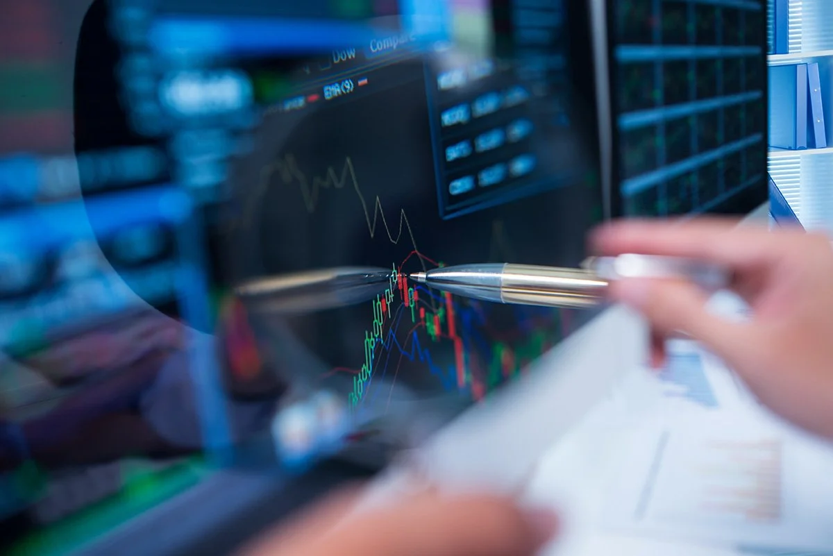 Close-up of financial charts on a computer screen with a person pointing at the graph using a silver pen.