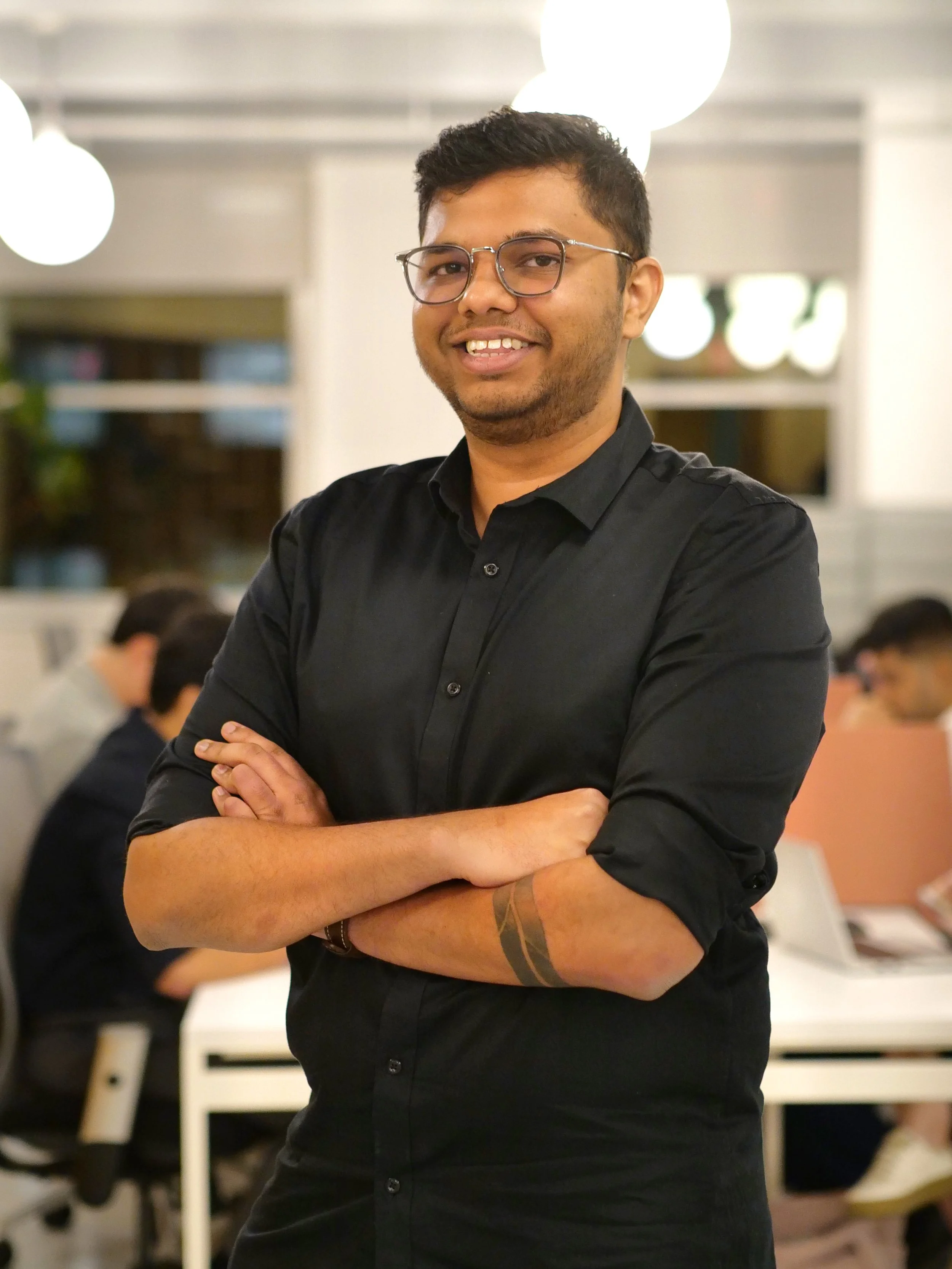 Headshot of a young man with dark hair wearing a suit and light blue collared shirt, standing indoors with a blurred modern building background.