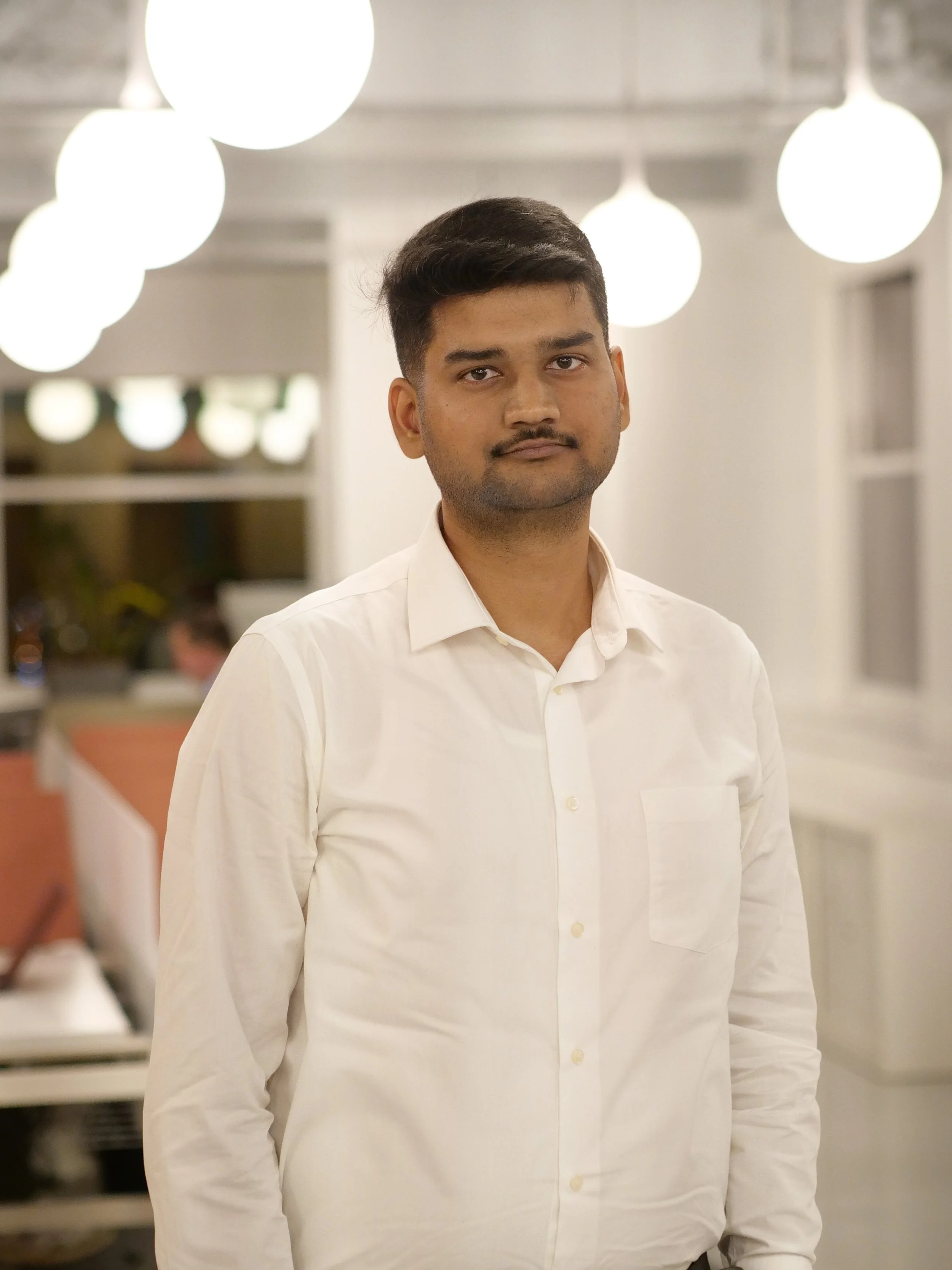 Professional portrait of a man in a dark suit and white shirt, standing in a modern, blurred office environment.