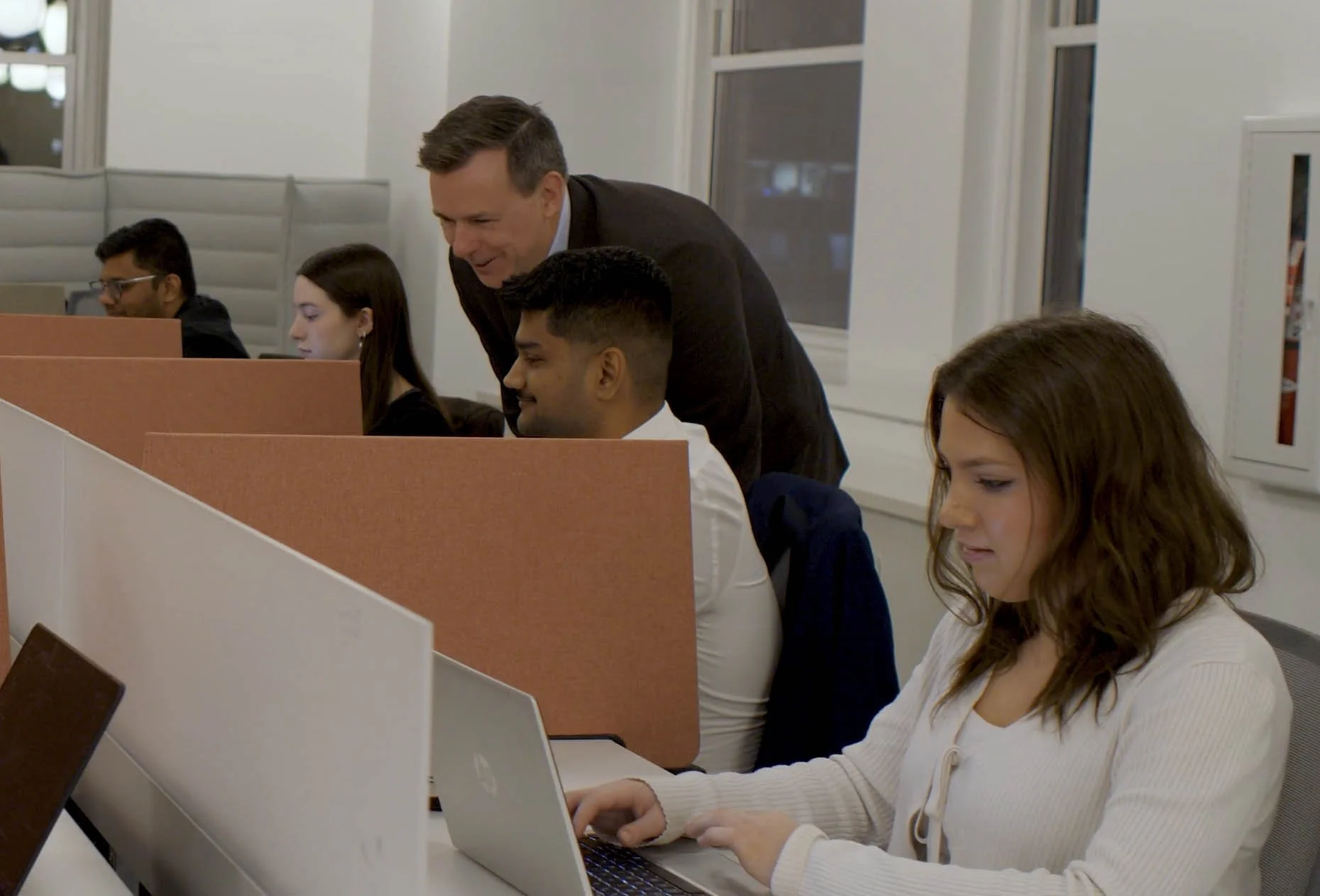 An office scene with diverse people working at desks with laptops, and a man standing and talking to a seated woman.
