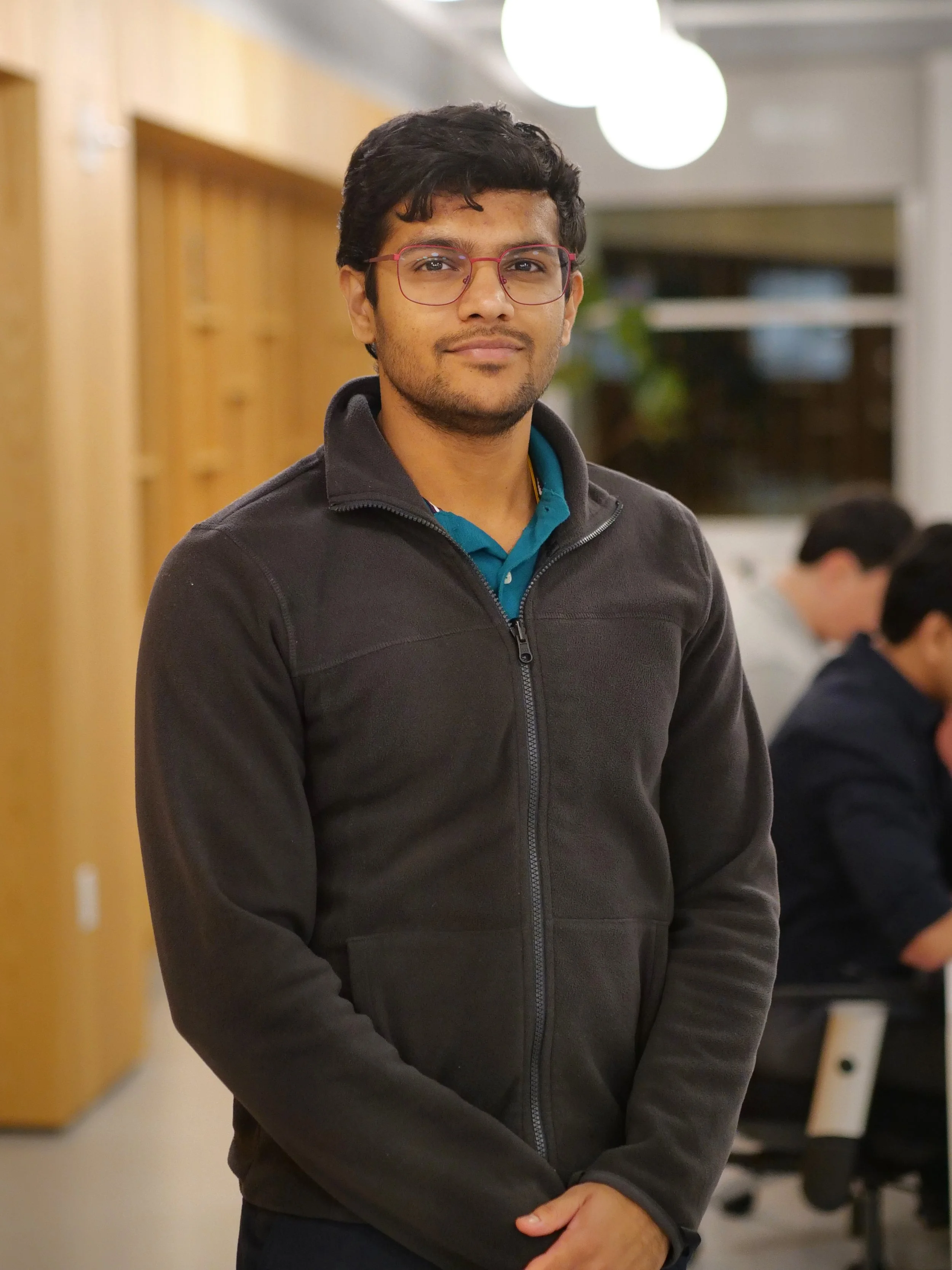 Professional portrait of a young man wearing glasses, a gray suit, a white shirt, and a red tie, smiling indoors.