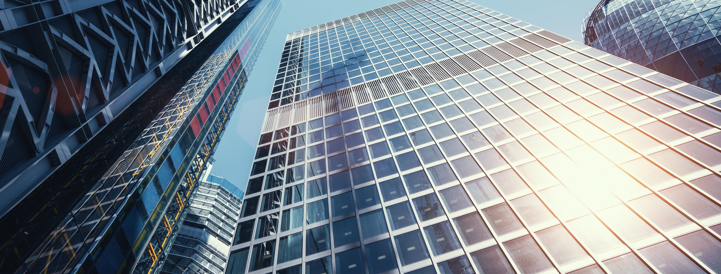 View of tall modern glass skyscrapers from street level, reflecting sunlight against a blue sky.