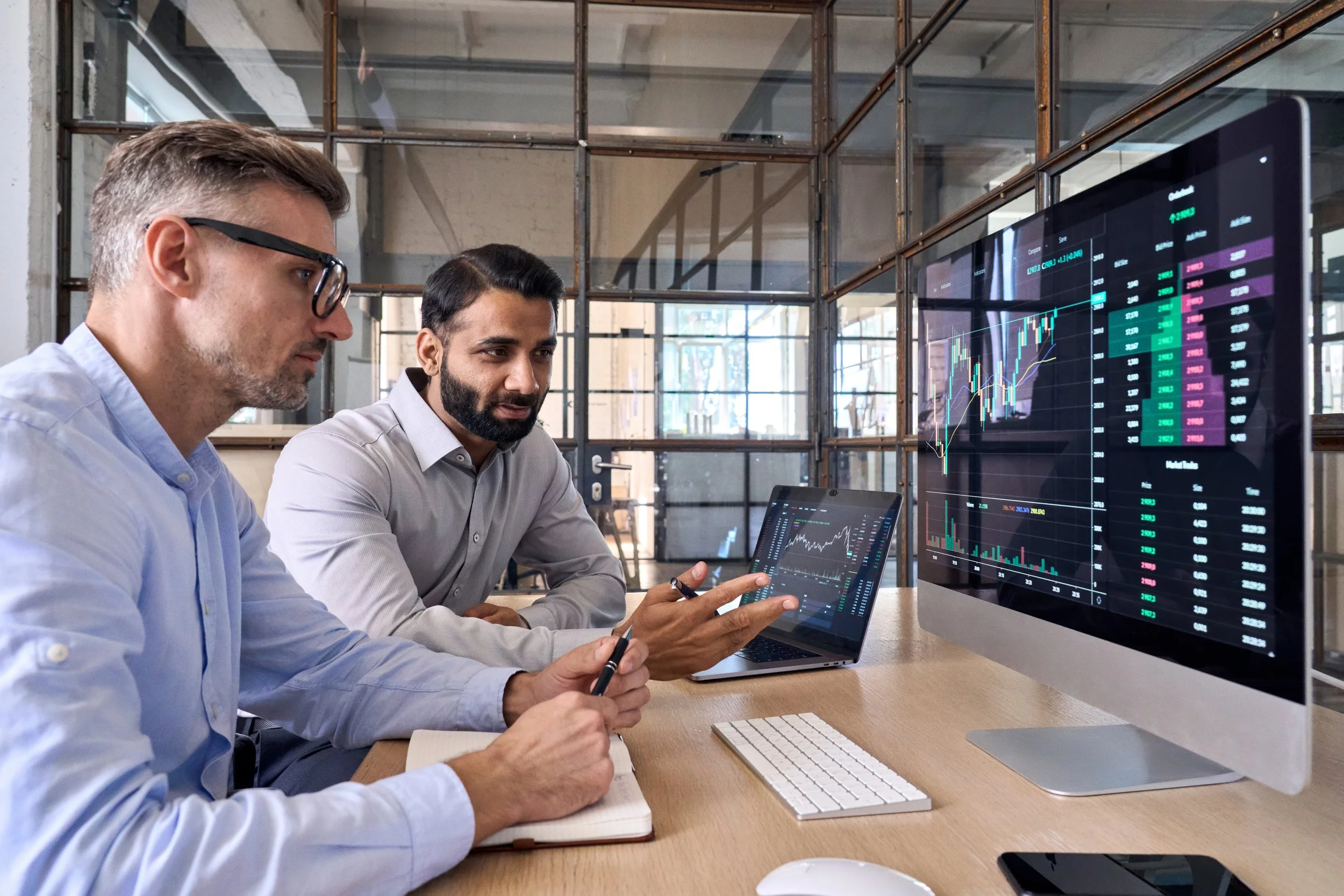 Two men sitting at a desk with multiple screens displaying stock market data and charts, discussing the information.