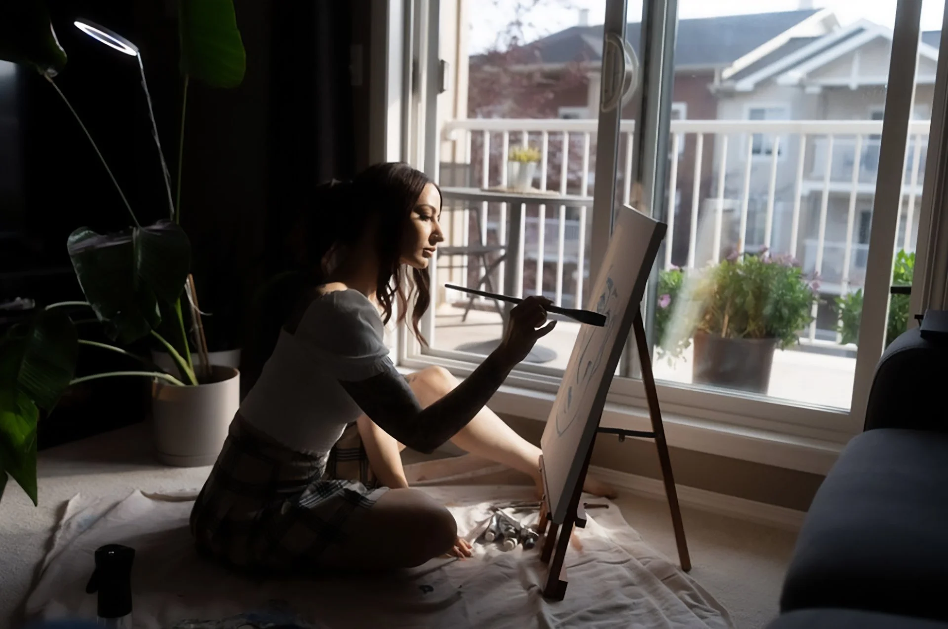 A woman sitting on the floor by a large window, painting on a canvas set up on an easel during daytime.
