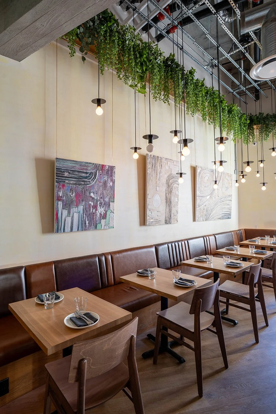 An interior of a modern restaurant with wooden tables, chairs, and a brown leather banquette. The wall features three abstract paintings, and hanging light fixtures with exposed bulbs are suspended from the ceiling, which is decorated with green plants and metal piping.
