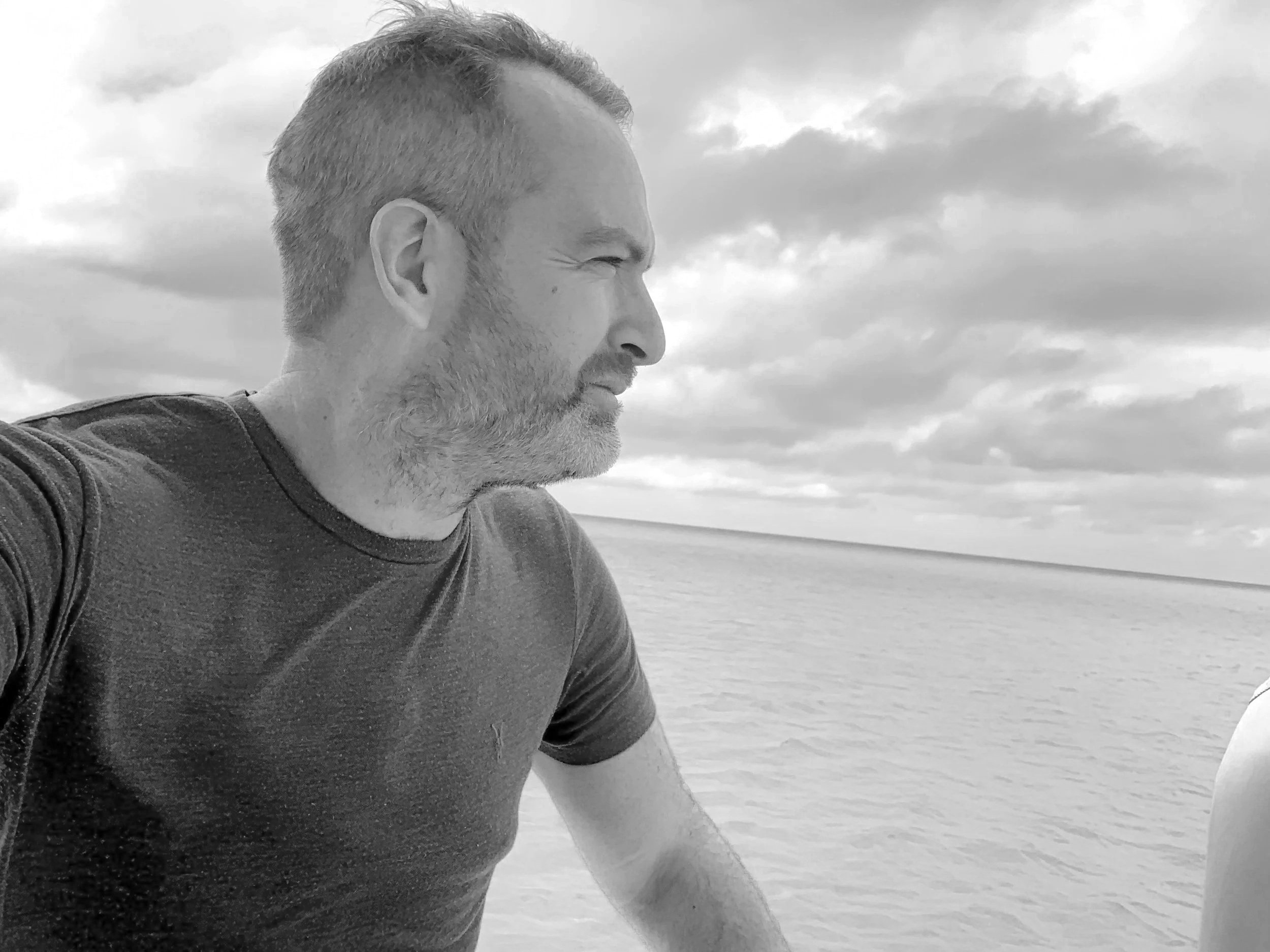 Andrew Zikking, Executive Producer. Or as AI puts it, simply... "a man with short hair and beard, wearing a T-shirt, looking to the right over a body of water with a cloudy sky in the background."