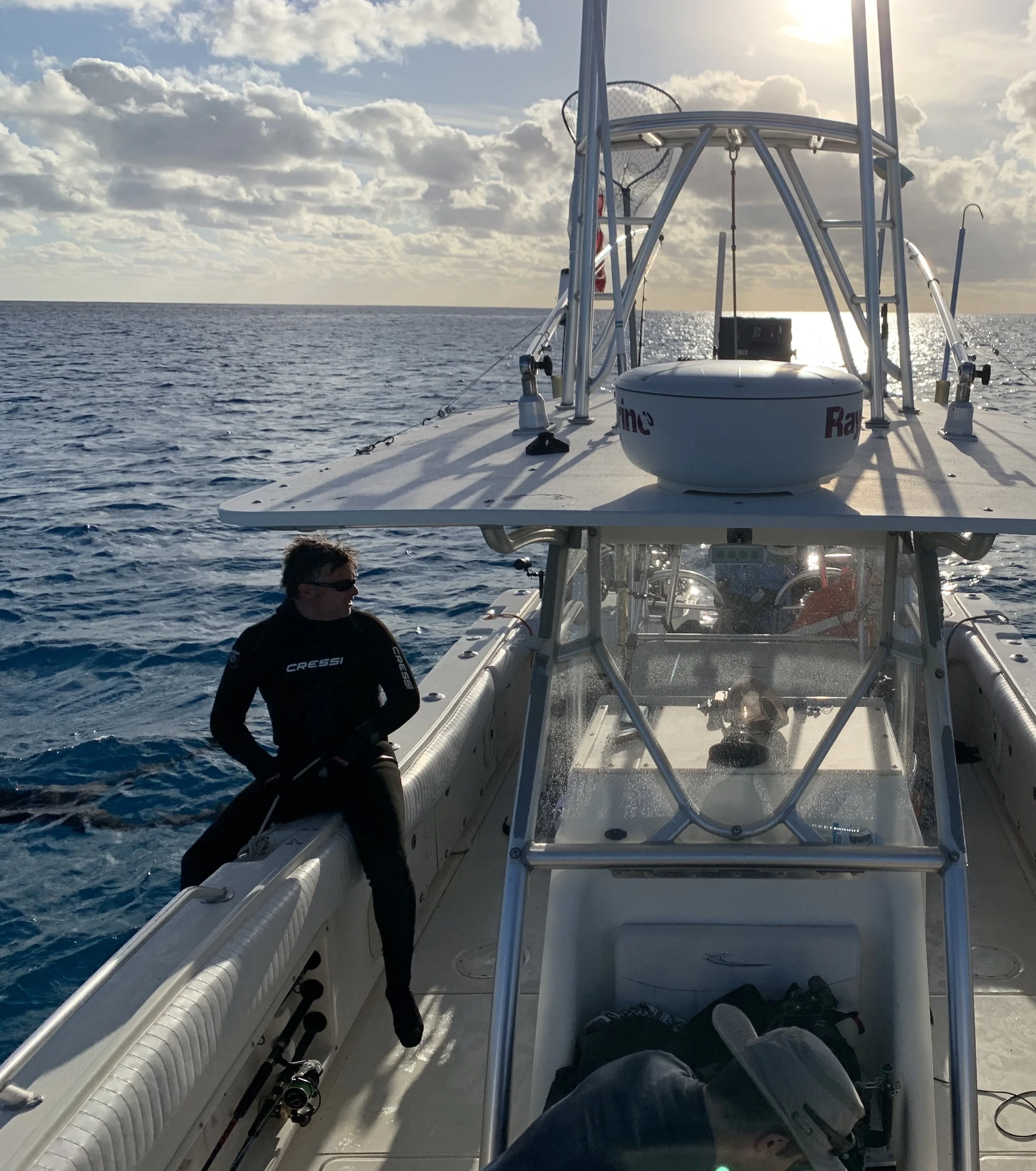 A person sitting on the side of a boat, wearing black, sunglasses, and a wetsuit, with the ocean and sky in the background.