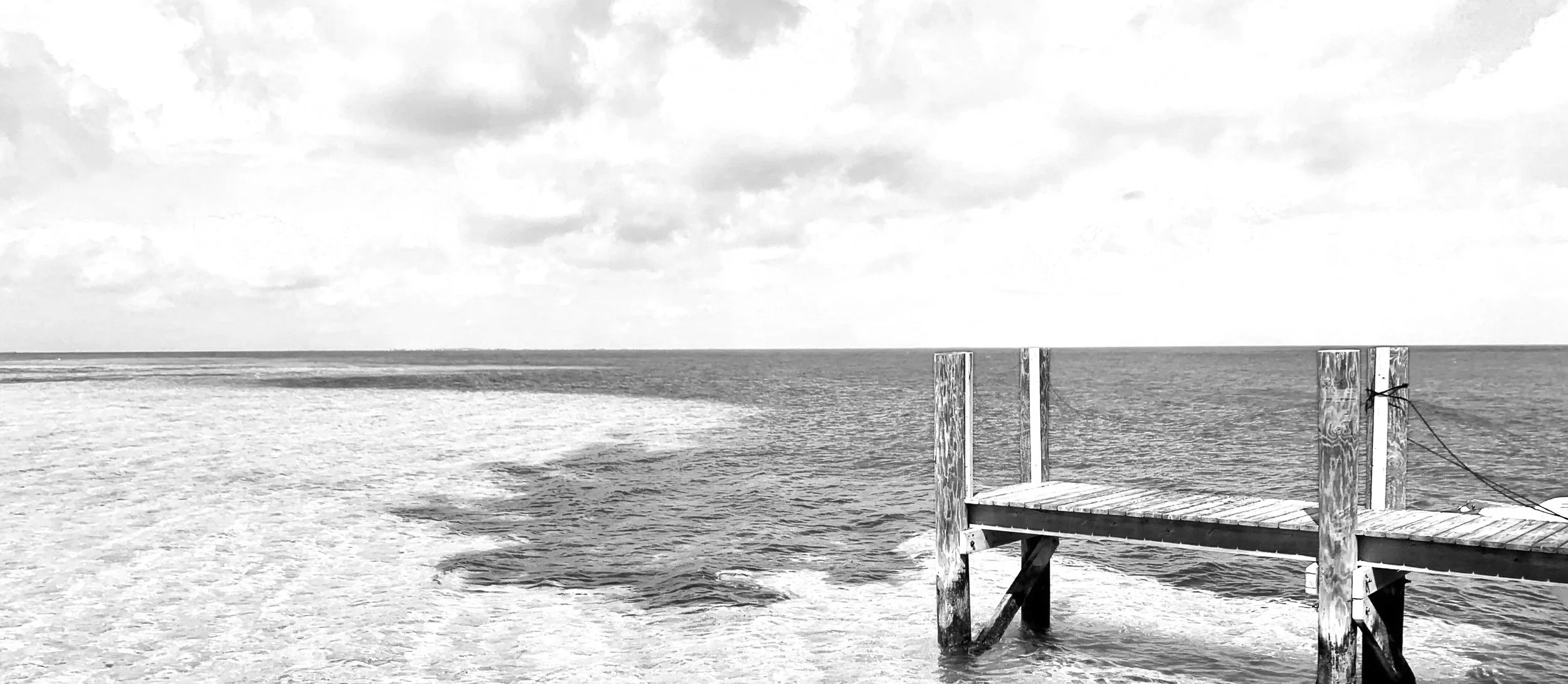 Bahamas dock. Black and white photo of a wooden dock extending into the water with boats nearby, under a partly cloudy sky.