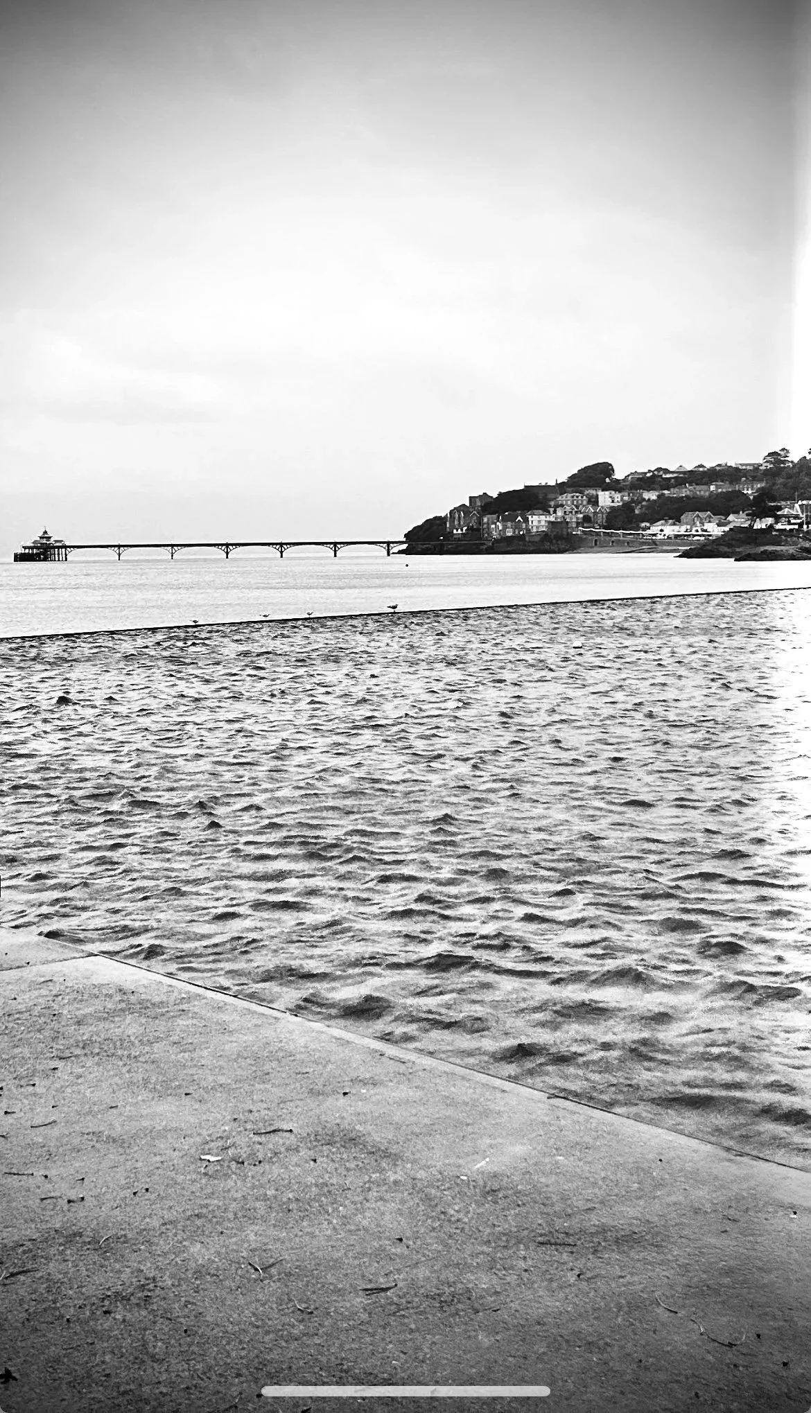 Clevedon Marine Lake. Black and white photo of a coastal landscape with water in the foreground, a pier extending into the water on the left, a bridge further out, and a hillside with houses in the background.