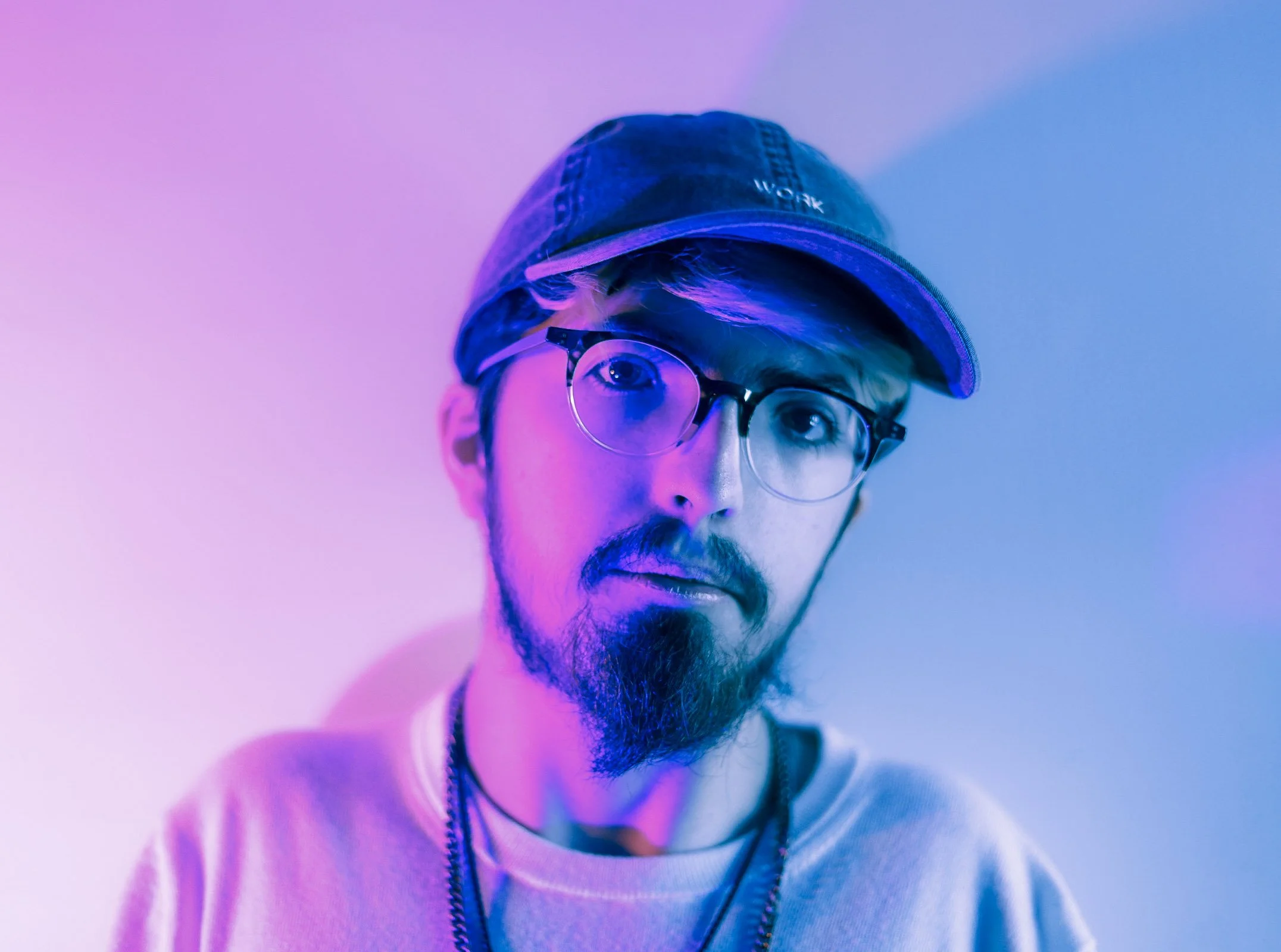 A young man with glasses and a beard wearing a cap and a light-colored shirt, illuminated by purple and blue lighting.