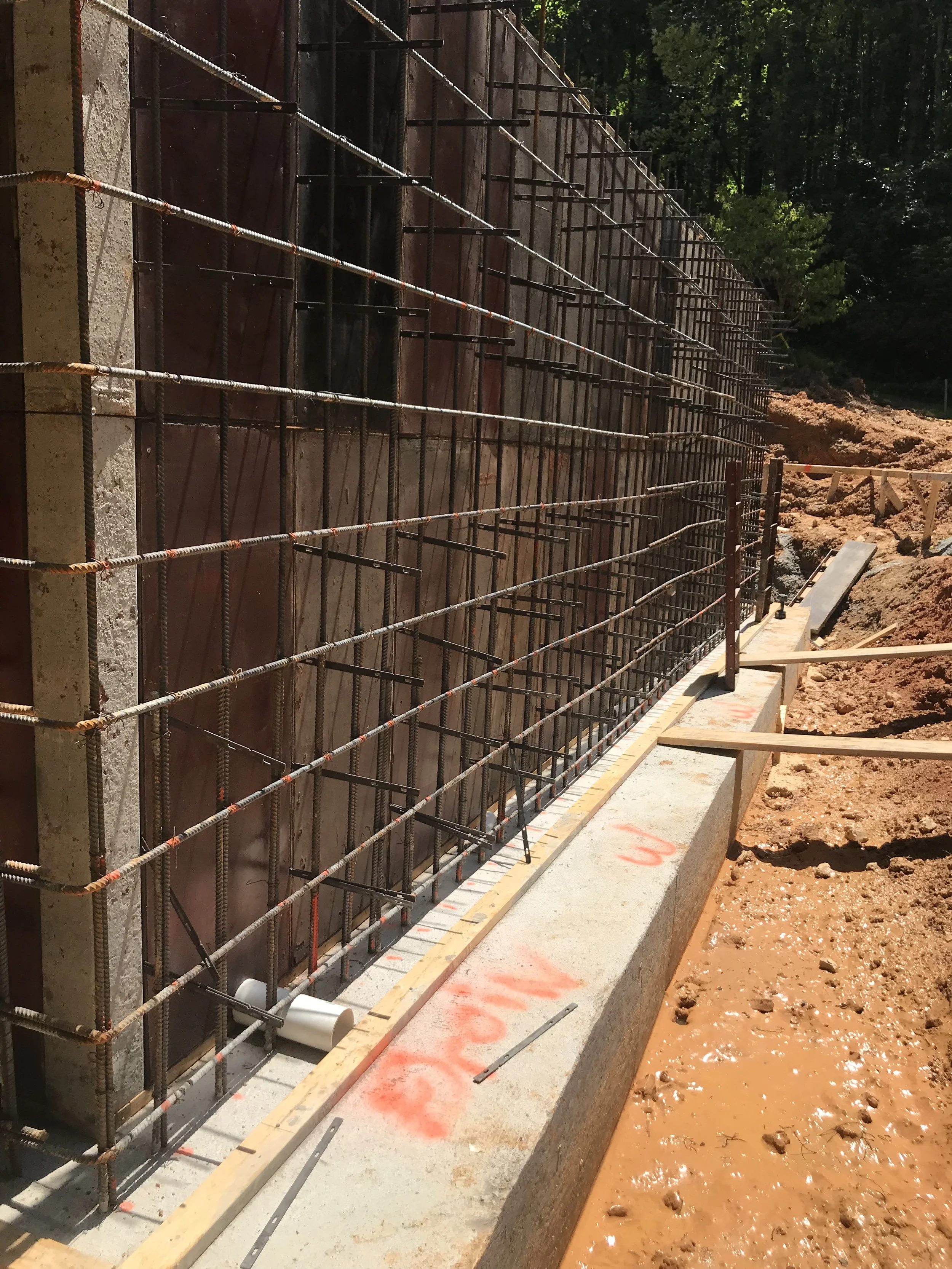 Construction site with rebar framework for a concrete wall, set into a muddy area with wood forms and construction markings.