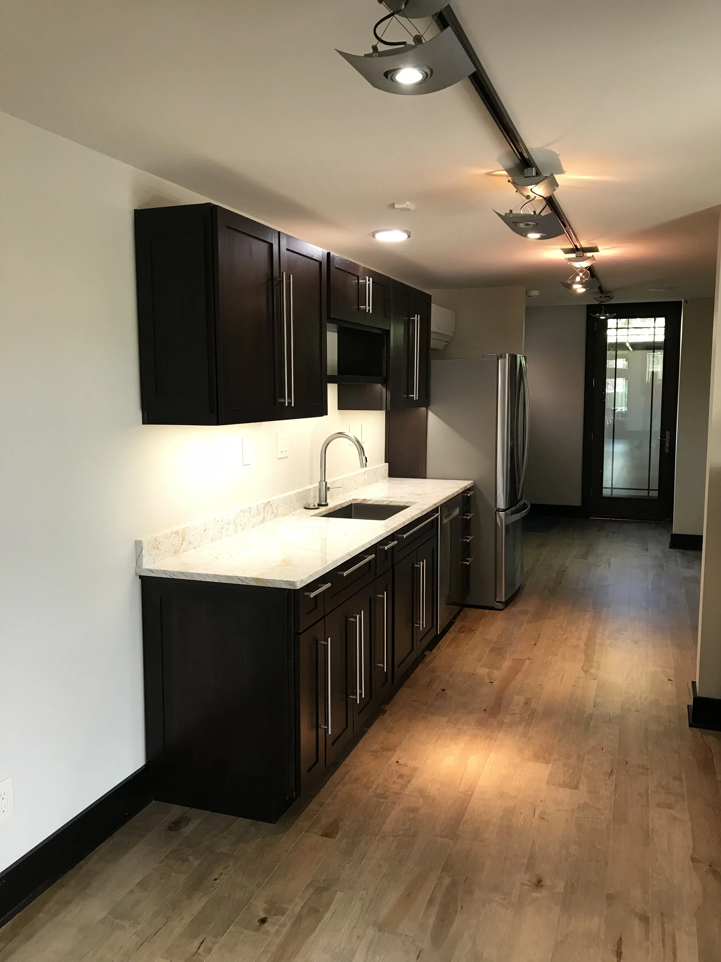 Modern kitchen with dark wood cabinets, white granite countertops, stainless steel refrigerator, and hardwood flooring.